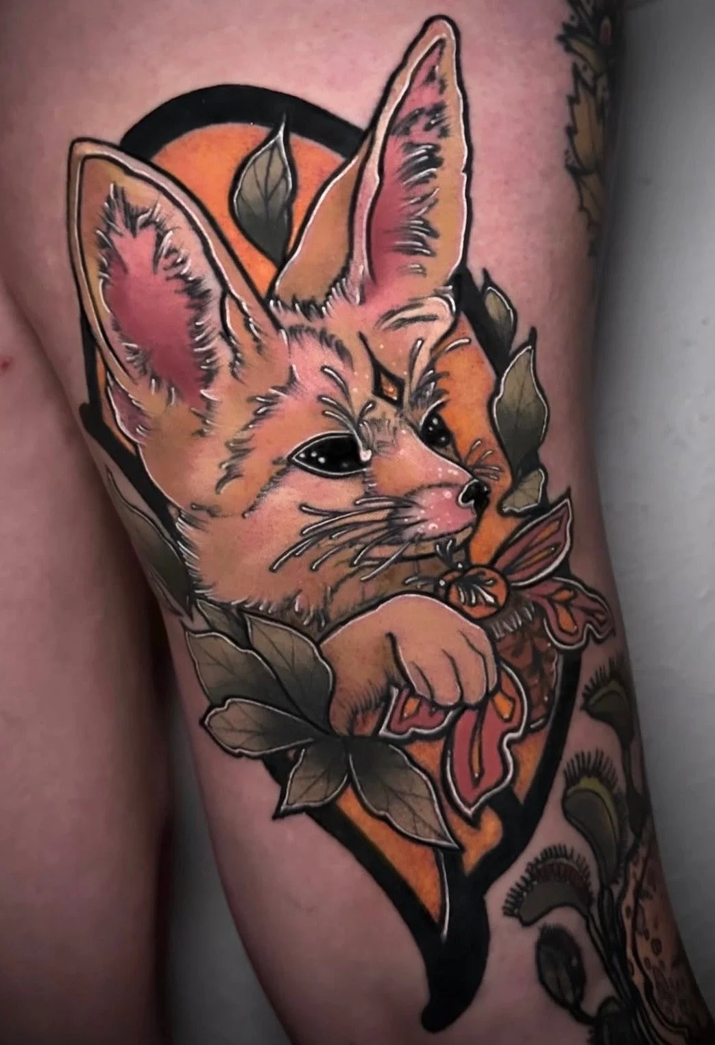 Colorful tattoo of a kitten with large eyes, surrounded by autumn leaves and a butterfly, with a bell in its paw.