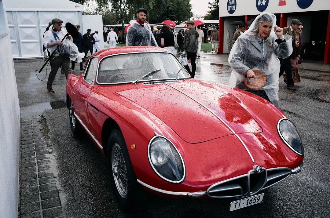 A red vintage Alfa Romeo sports car with raindrops on its surface, parked at an outdoor event with people walking around in raincoats and umbrellas.