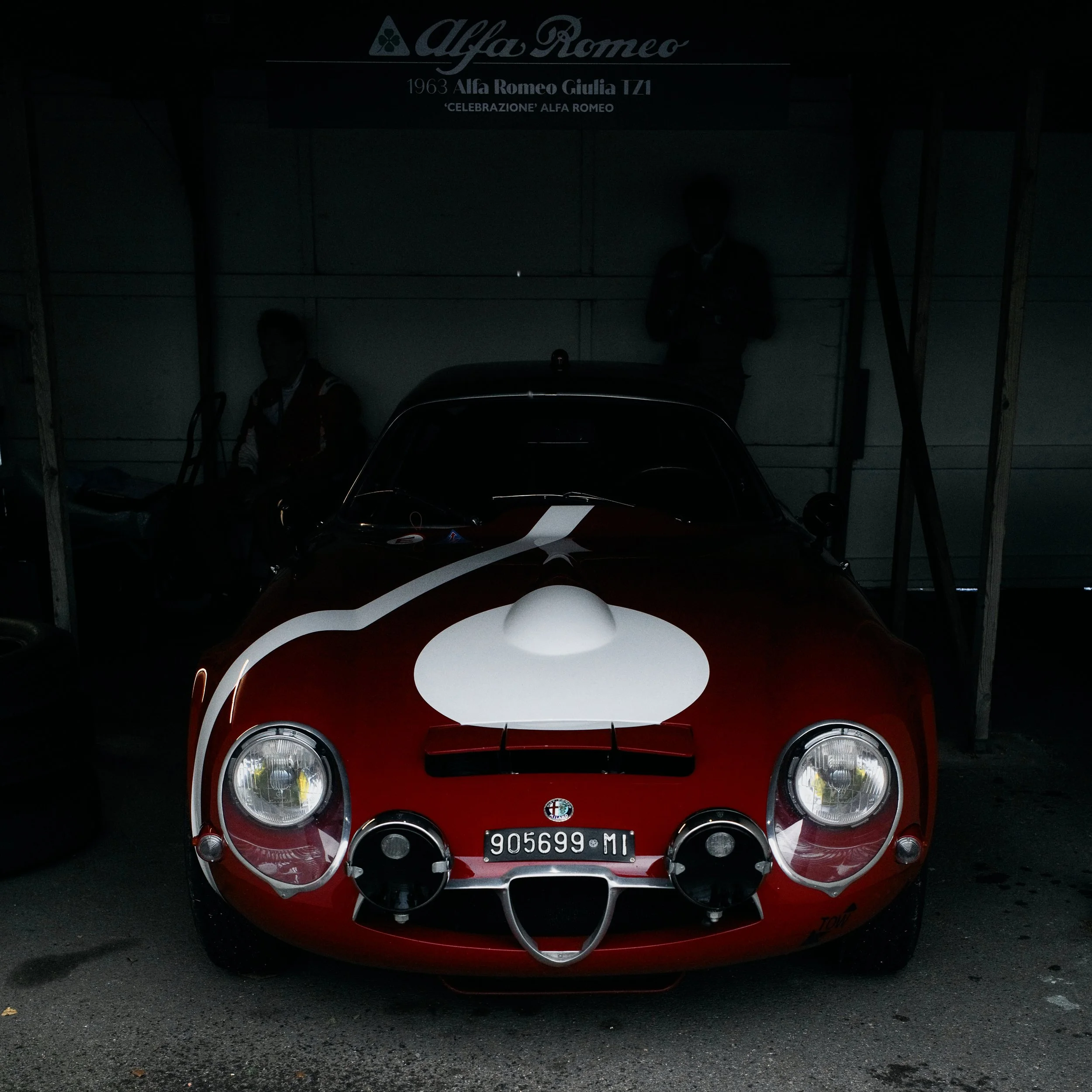 Red vintage Alfa Romeo race car with white racing stripe and Italian license plate, parked indoors with shadowed people in background.
