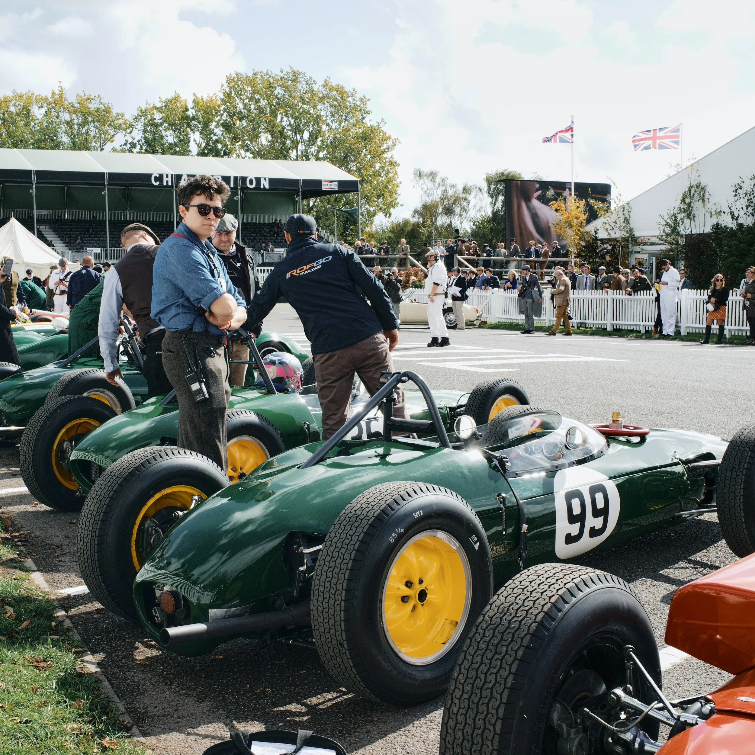 Green vintage race cars with yellow wheels at a racing event, with people around and a grandstand in the background.