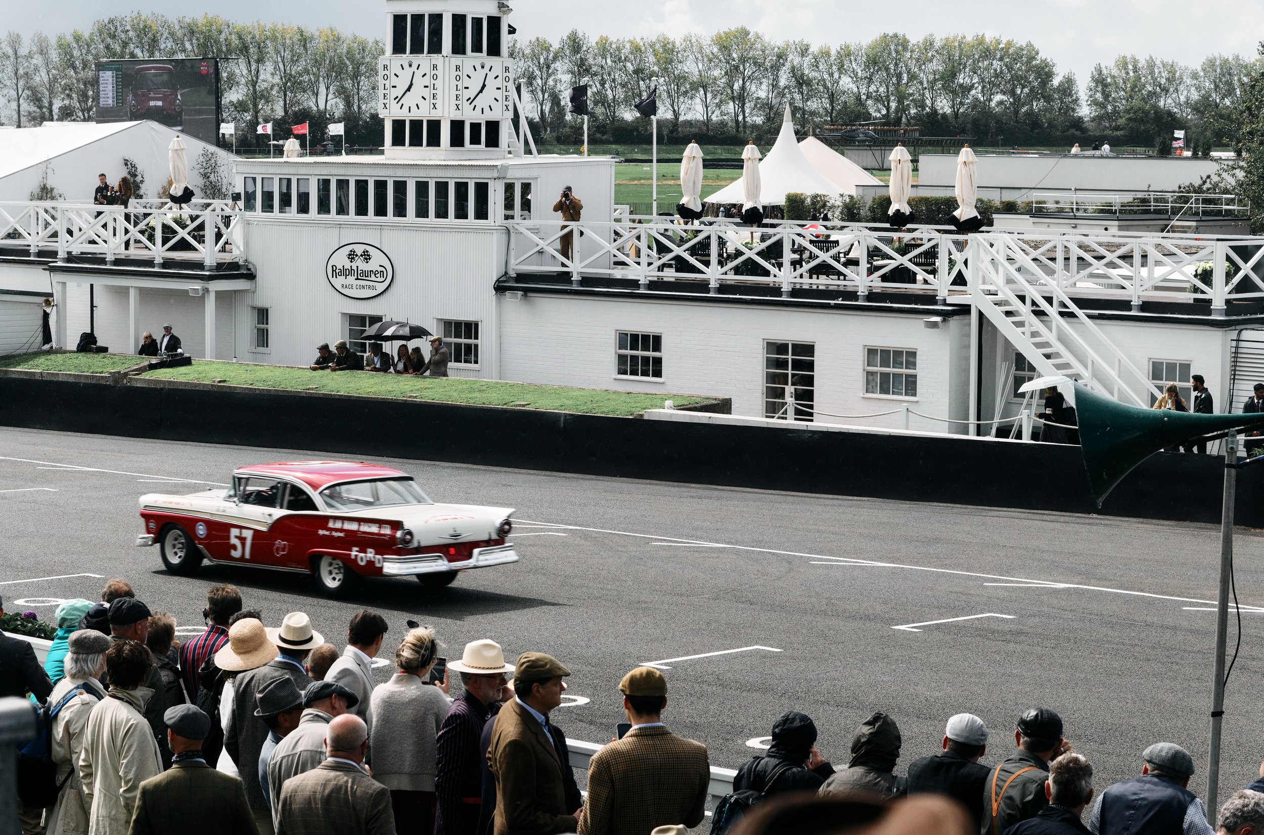Vintage race car driving past spectators at the Goodwood Revival motorsport festival with a white race control building branded Ralph Lauren Race Control, a scoreboard, and spectators and staff watching from the stands and ground level.