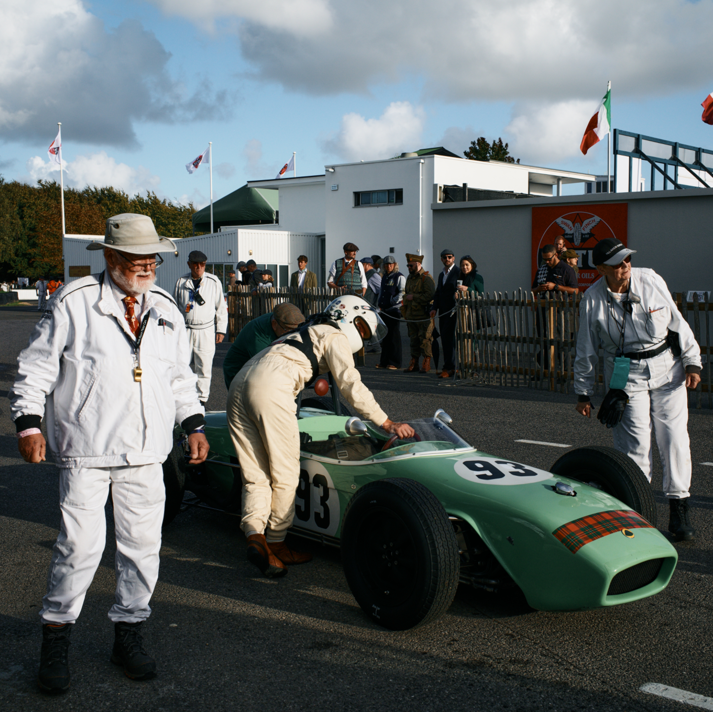A vintage green race car with the number 93, surrounded by people including a driver in a white racing suit and helmet. Several spectators and officials are observing the scene, with flags flying in the background and a building with signs nearby.