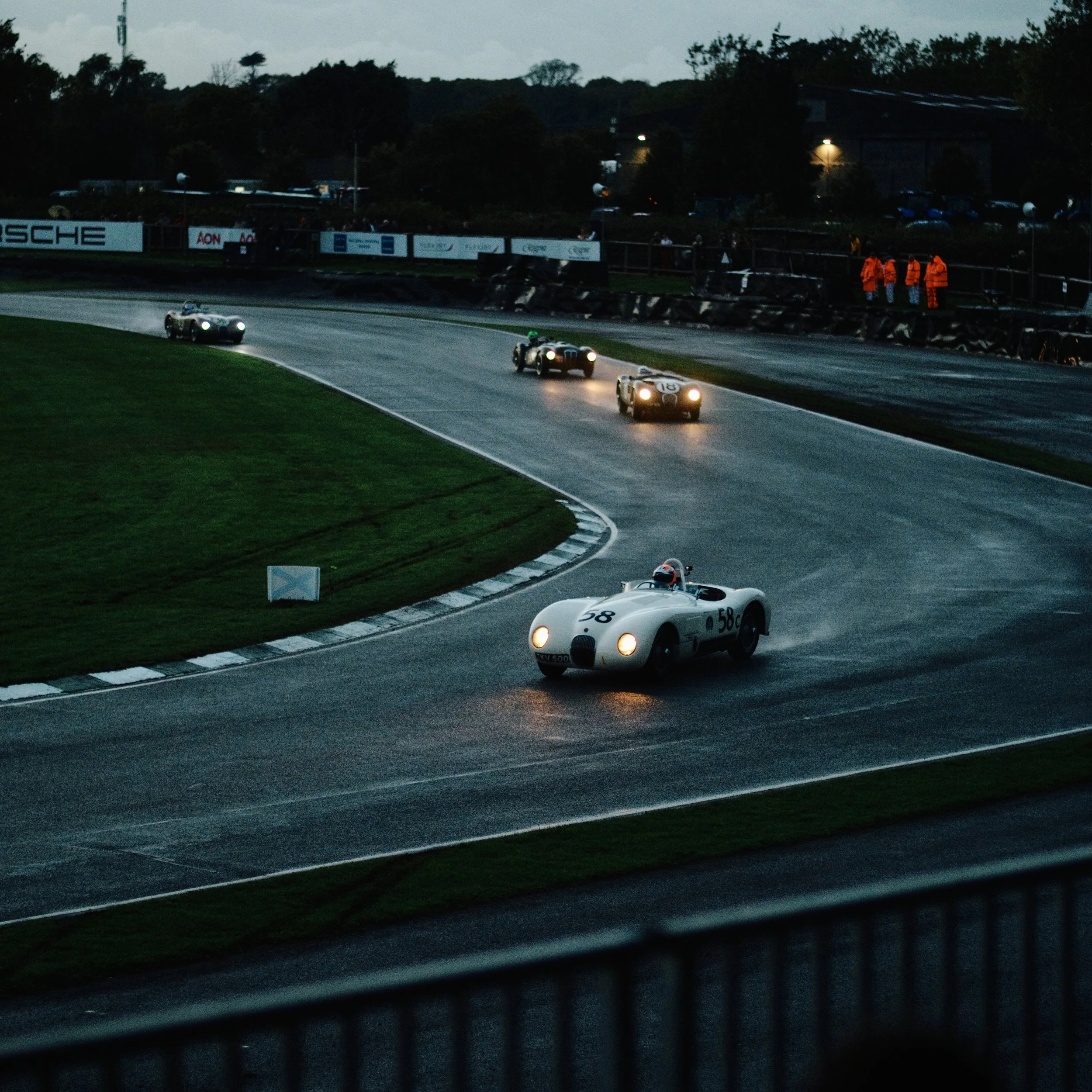 Vintage race cars driving on a wet track during dusk or evening, with a few spectators in bright orange jackets watching from the side.