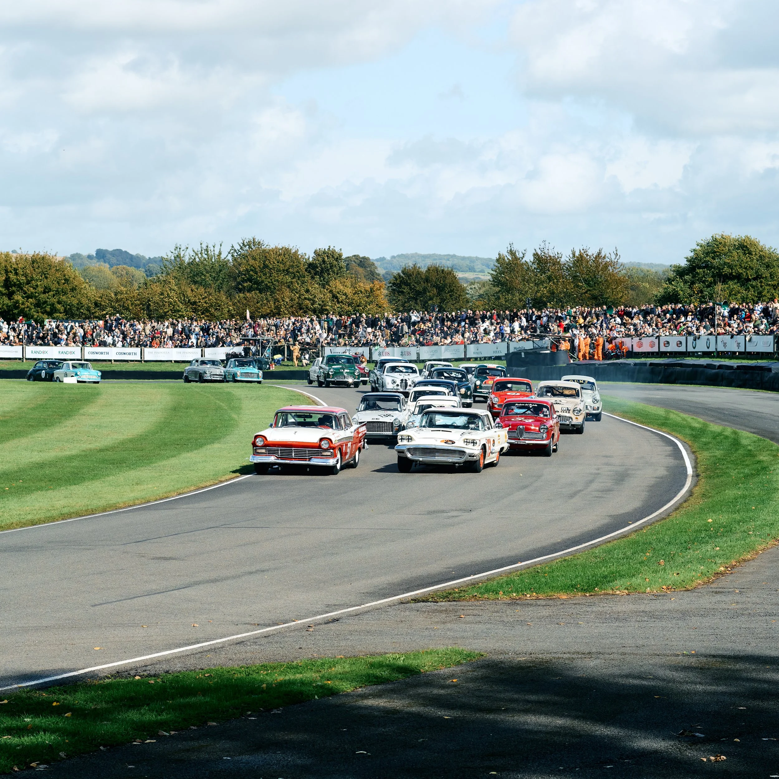 Multiple vintage race cars driving on a race track with a crowd watching from the sidelines and trees in the background.