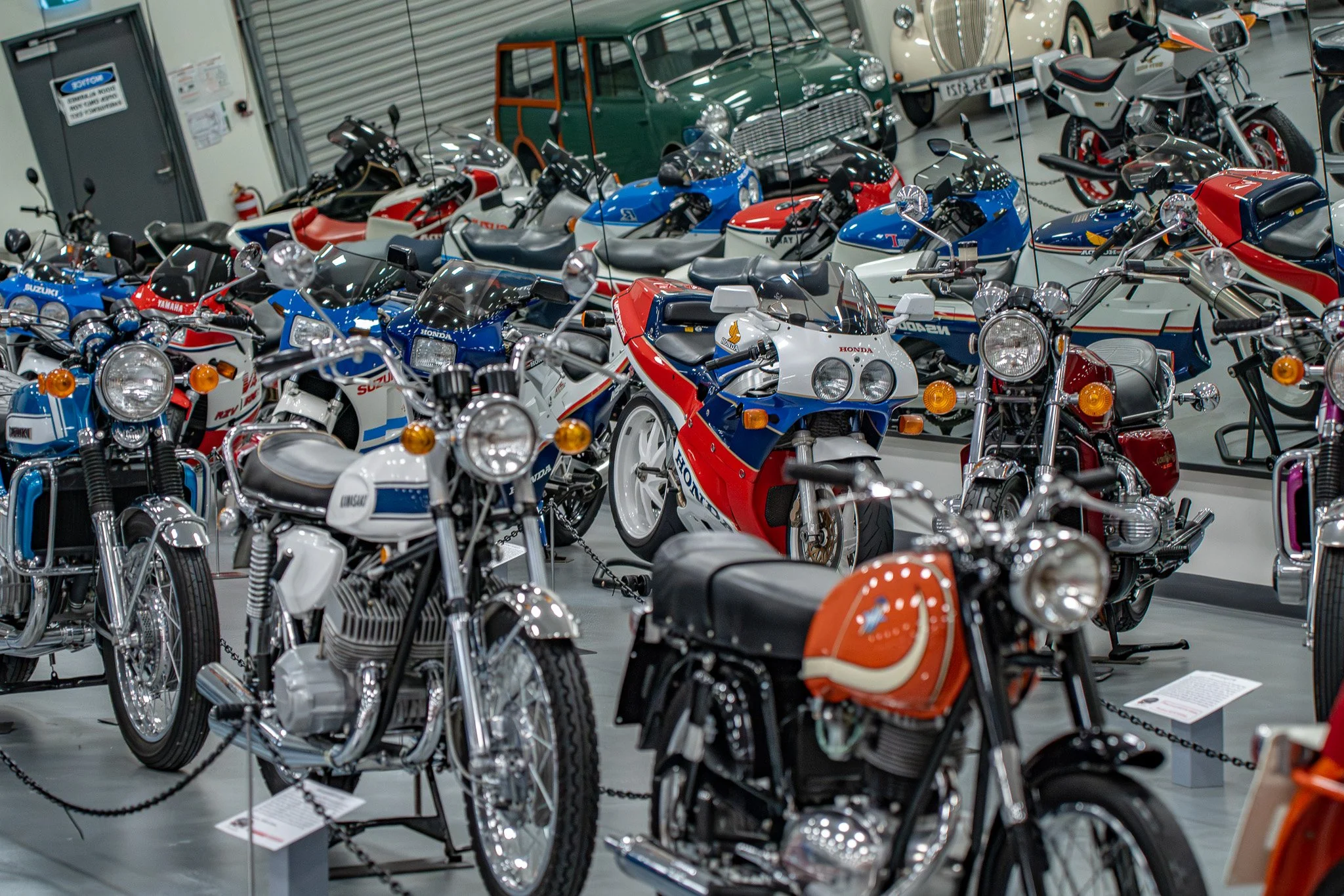 Several vintage motorcycles and a few classic cars are displayed in a showroom or museum, behind glass.