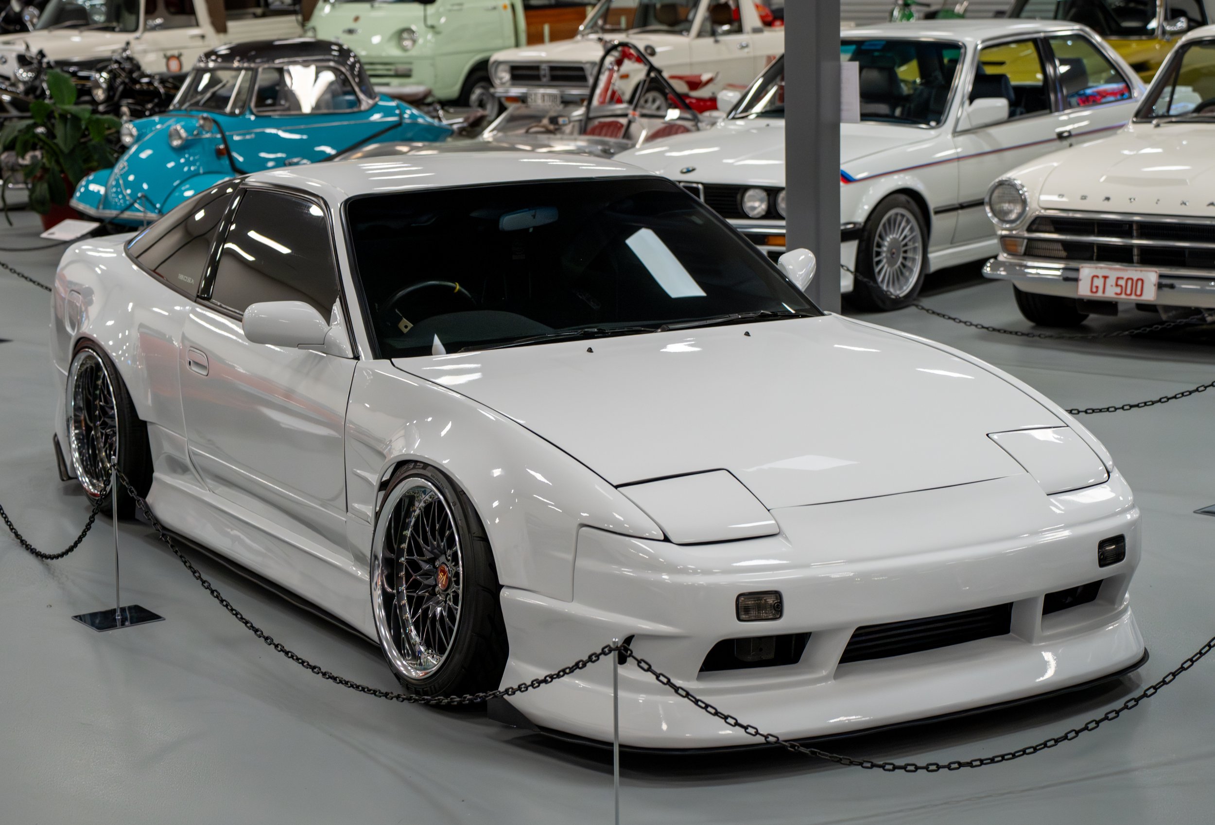 White sports car with black roof behind a barrier in an indoor car museum, other vintage cars and a blue vehicle are visible in the background.