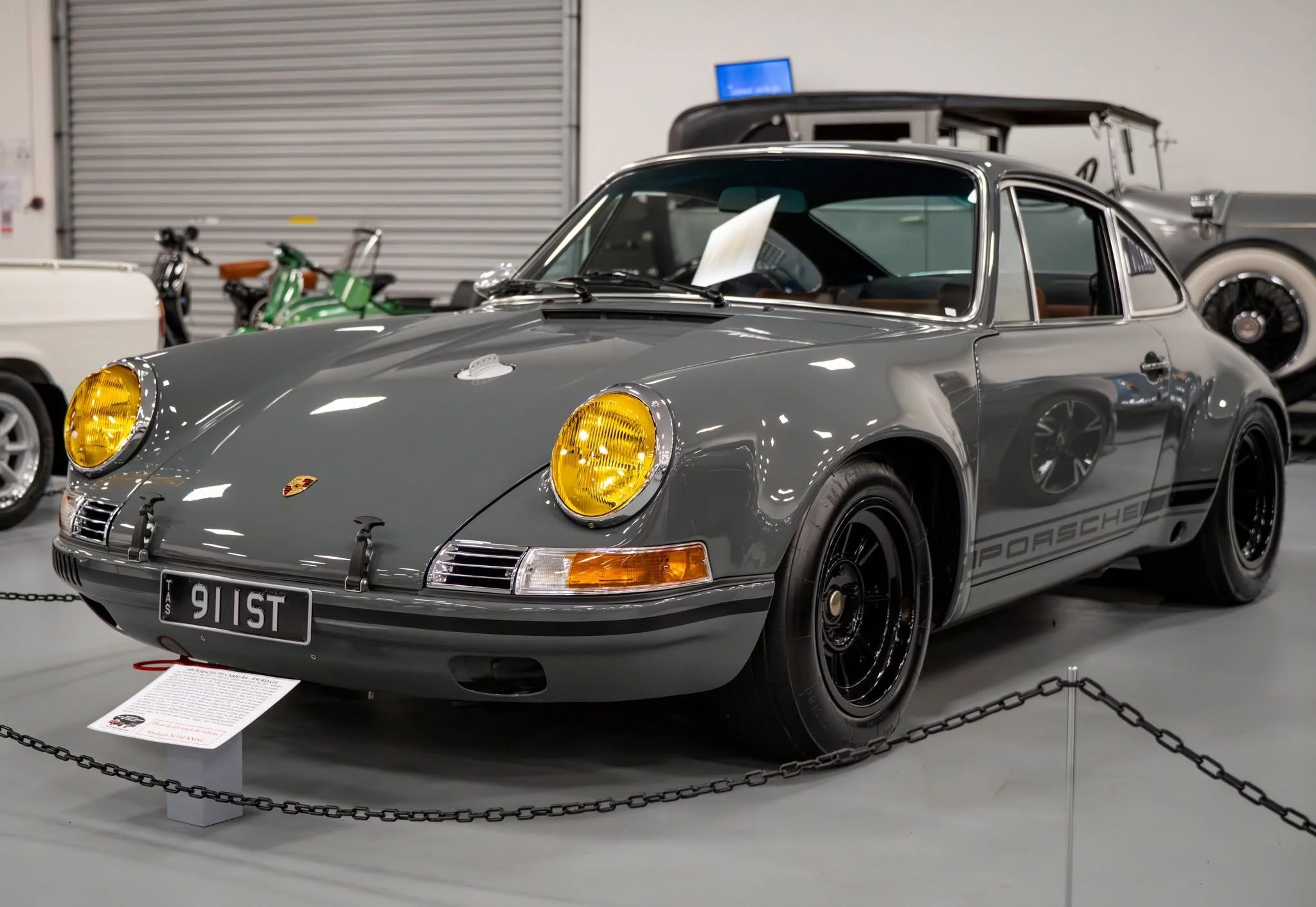 A vintage silver Porsche 911 with yellow headlights is displayed at a car show, surrounded by a black chain barrier and informational placard on the floor.