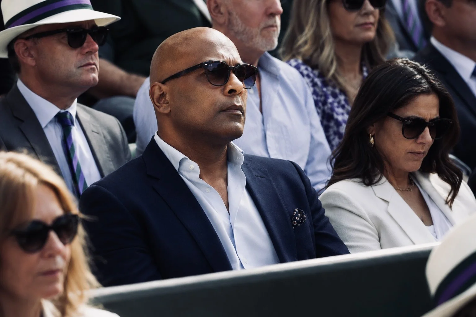 Roberto Revilla seated courtside at Wimbledon wearing a navy blazer, light blue open-neck shirt and gradient tint sunglasses, watching the tennis among a stylishly dressed summer crowd in bright sunshine.