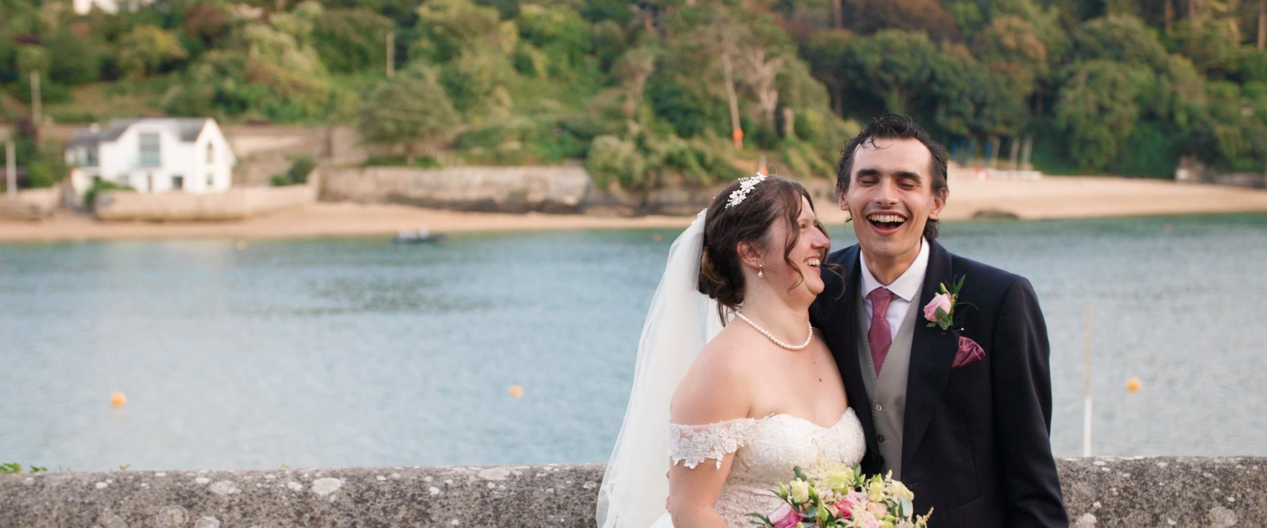 Amy&amp; Stephen laugh fiercly together as they stand in front of the Salcombe Estuary in their wedding outfits