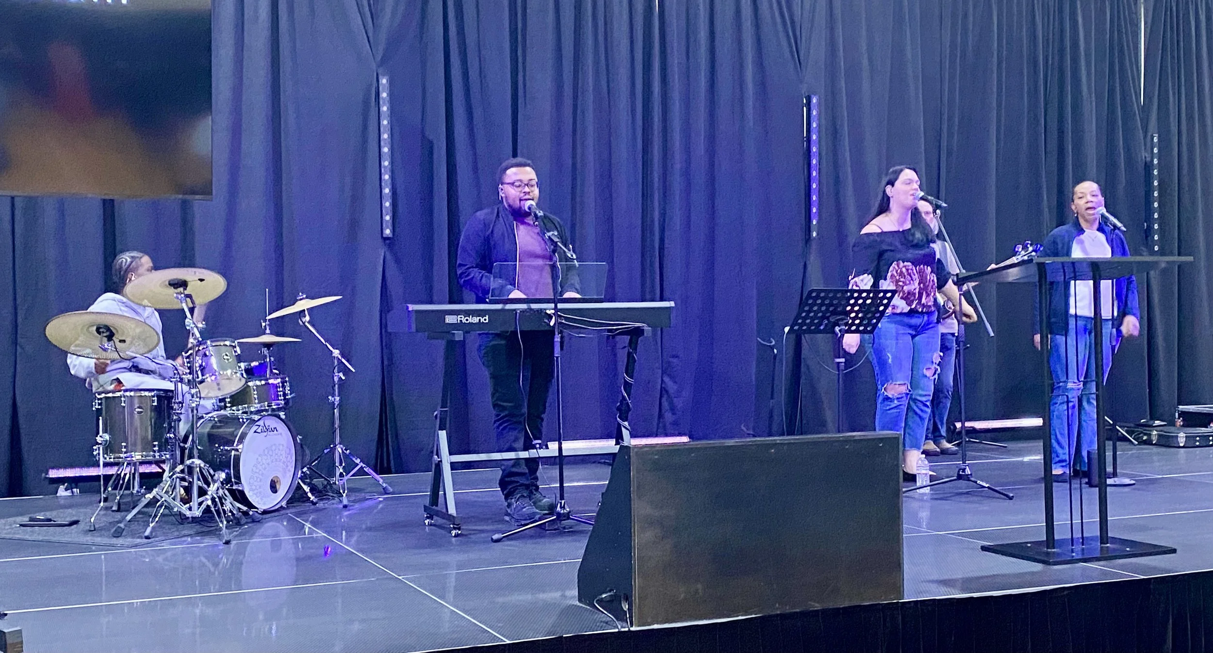 A musical performance on stage with four singers, a keyboard player, and a drummer, all singing into microphones, with a dark backdrop and stage lighting.