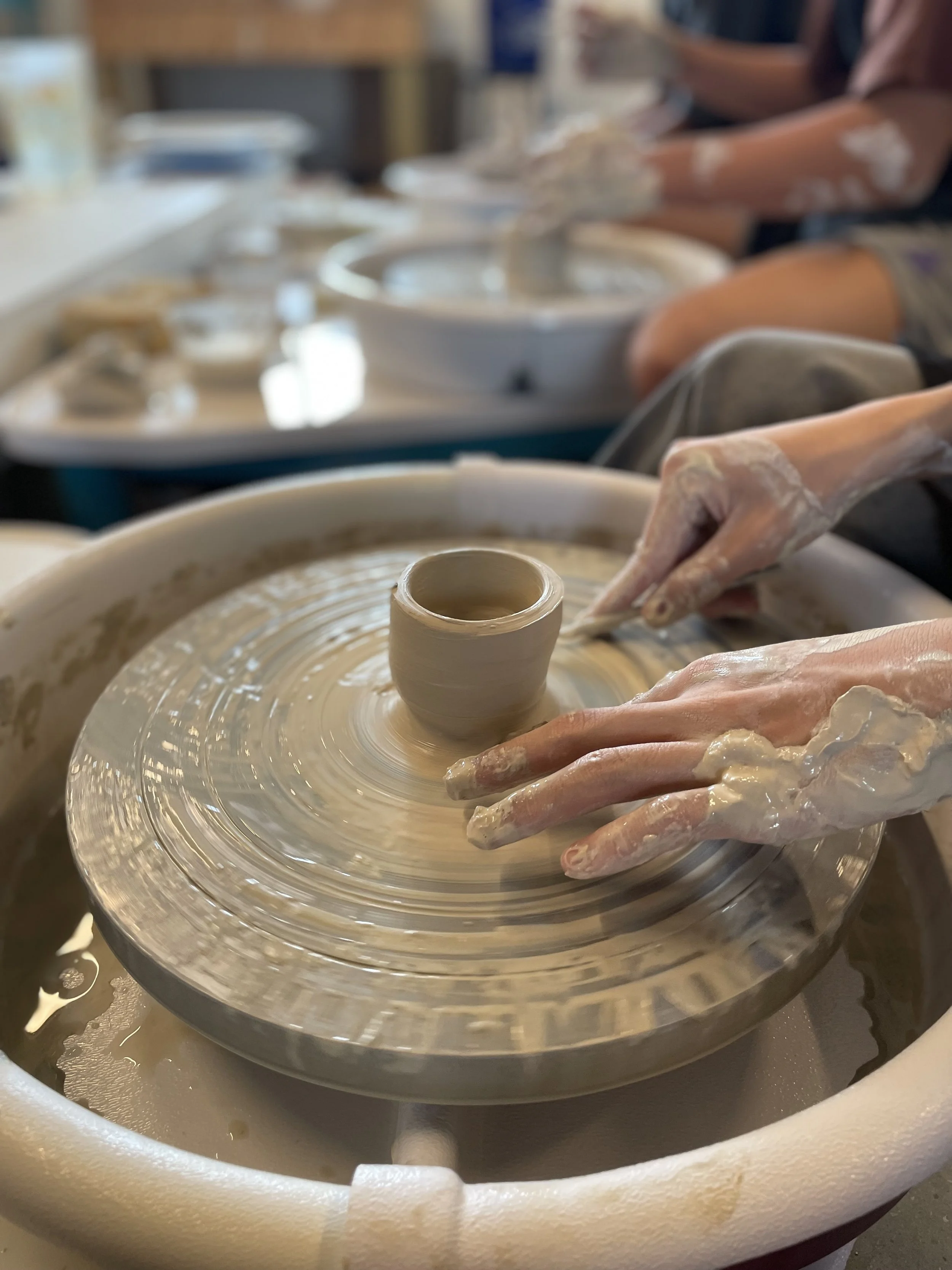 a set of hands sculpting clay on a ceramic wheel with a second pair of student hands holding sponge
