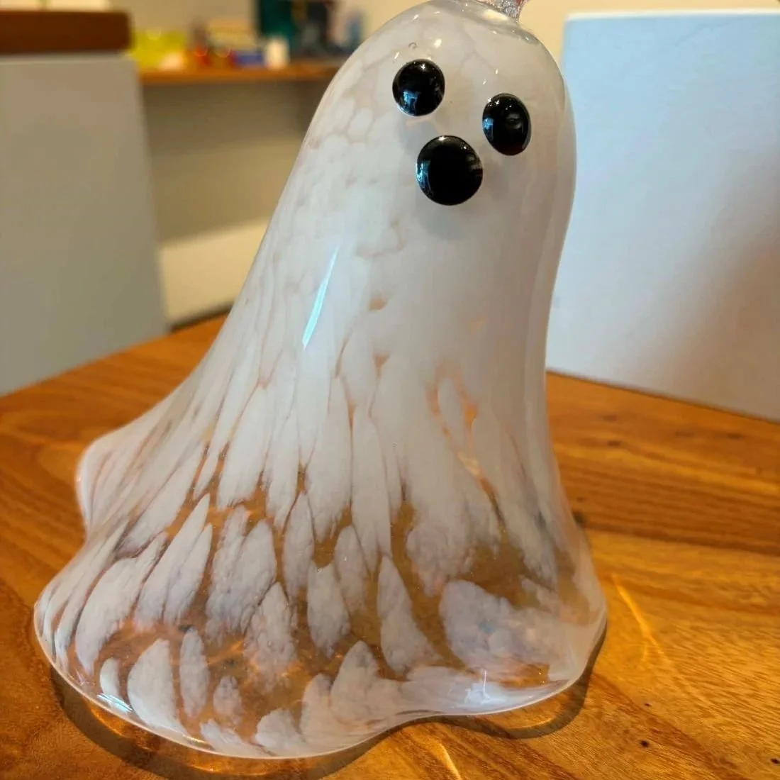 a glass blown white ghost with cute black eyes and nosesits in a blurry art shop