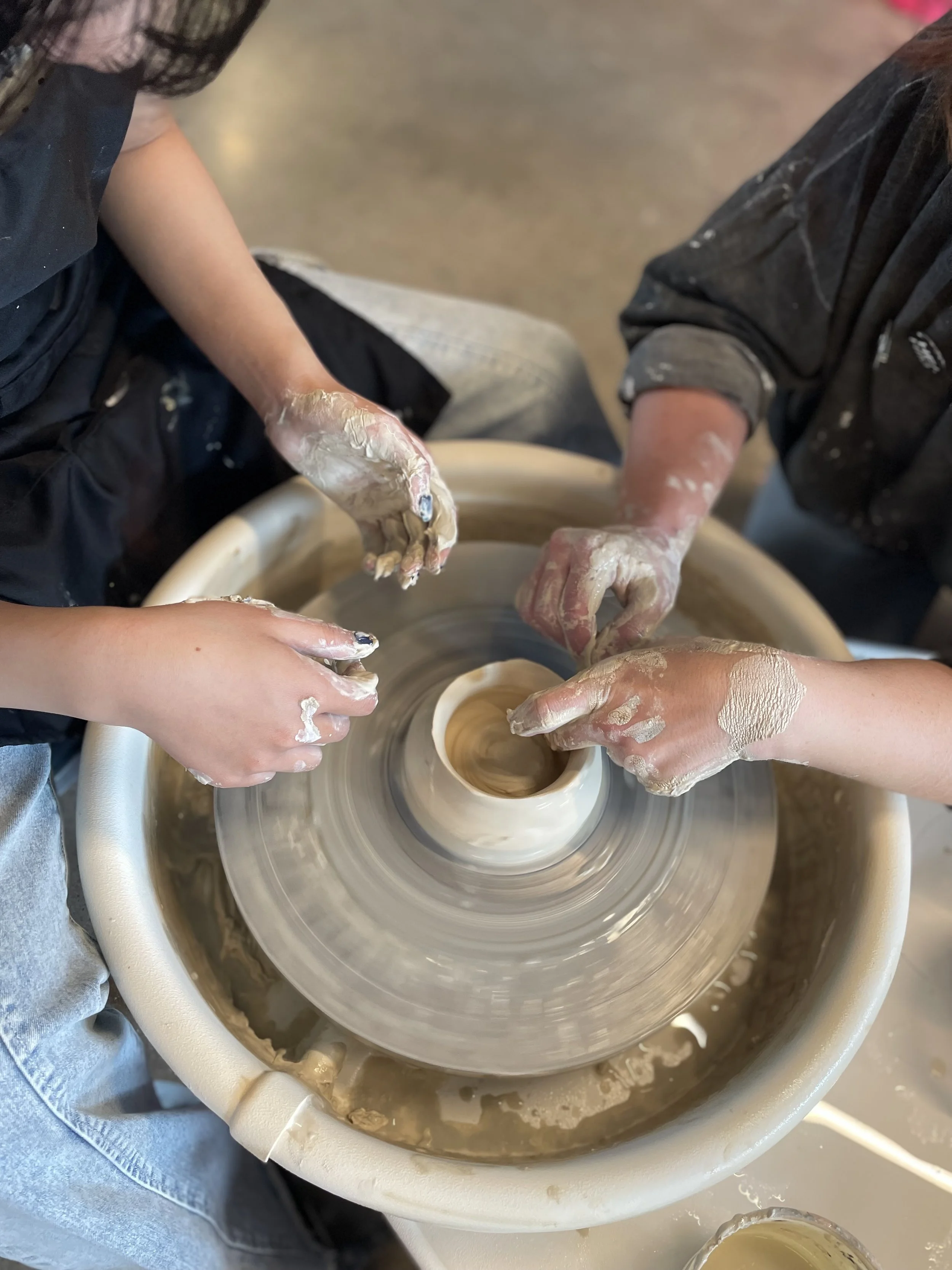 a set of hands sculpting clay on a ceramic wheel with a second pair of student hands holding sponge