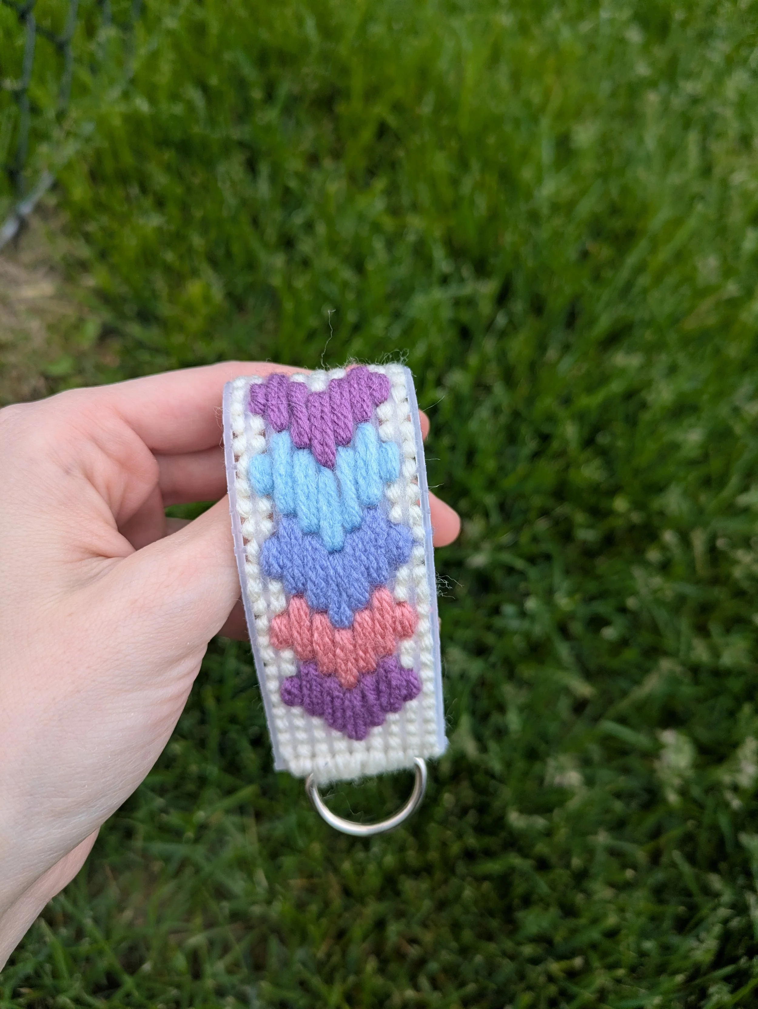 a white needlepoint keychain with multi colored hearts on a grassy background