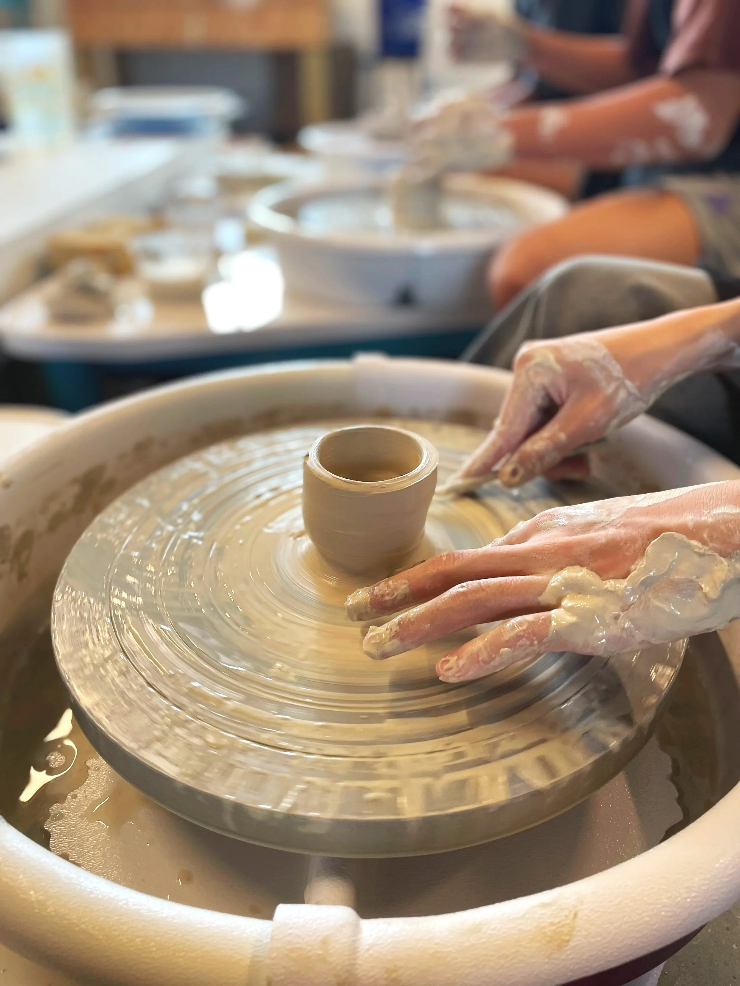 a set of hands sculpting clay on a ceramic wheel with a second pair of student hands holding sponge
