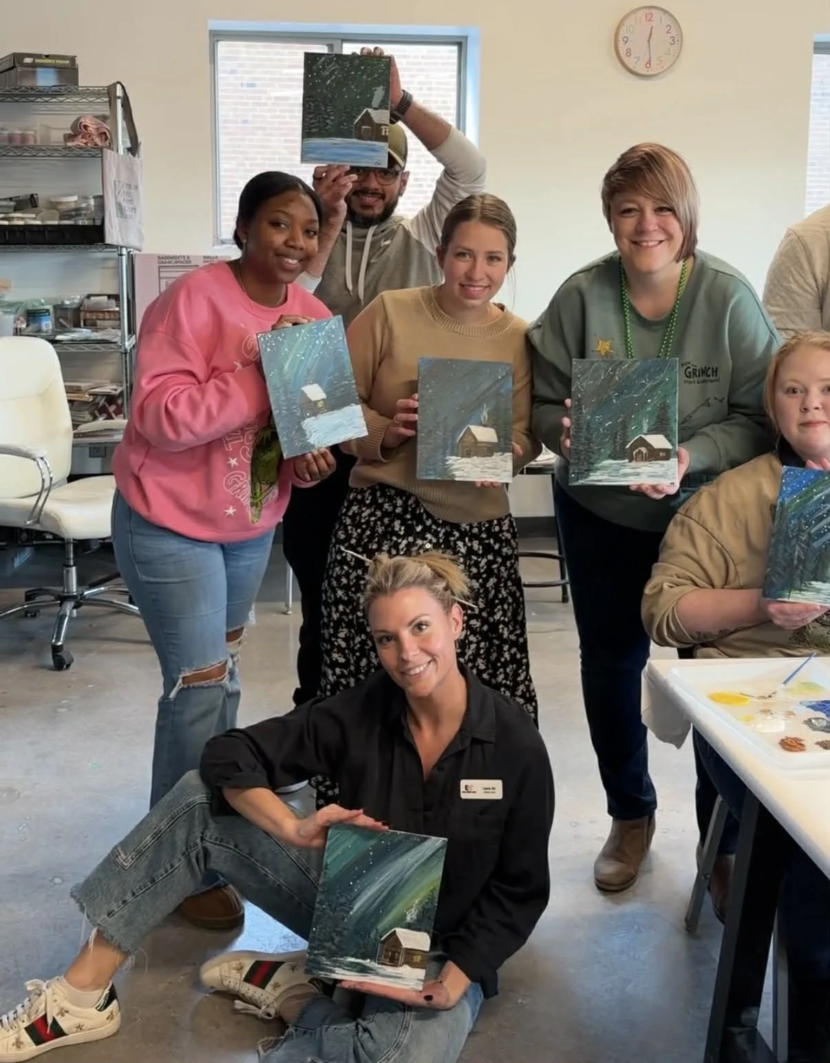 A group of people pose with their completed paintings of a snowy cabin scene.