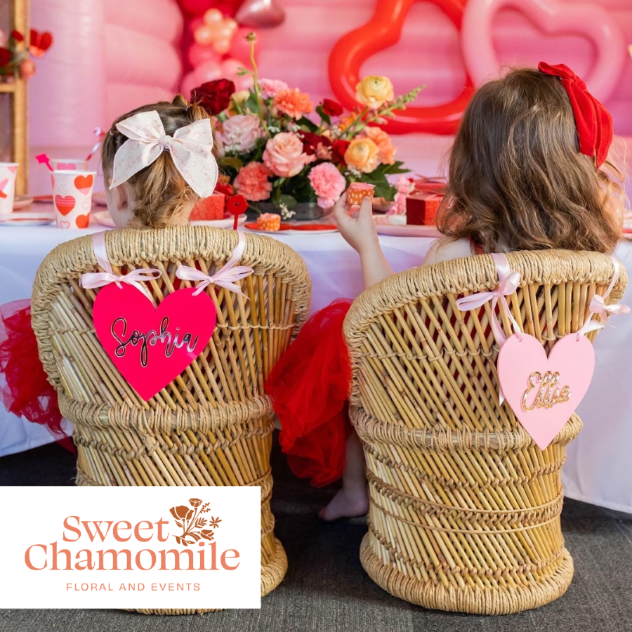 Two girls in wicker chairs with heart decorations looking at a Valentines Day floral arrangement.
