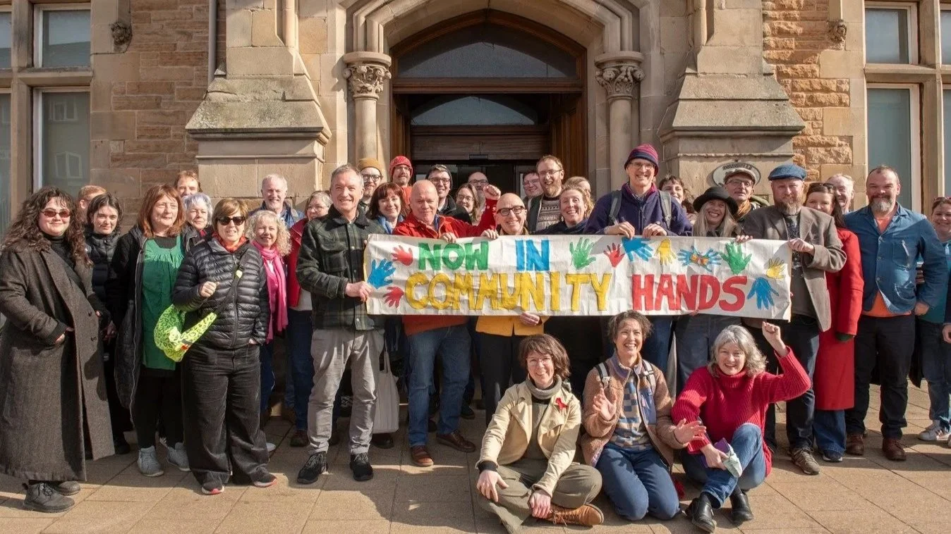 A group of people smiling, standing in front of the door of the former Portobello police station, holding up a homemade banner with colourful writing that says 'NOW IN COMMUNITY HANDS' with colourful handprints on it.