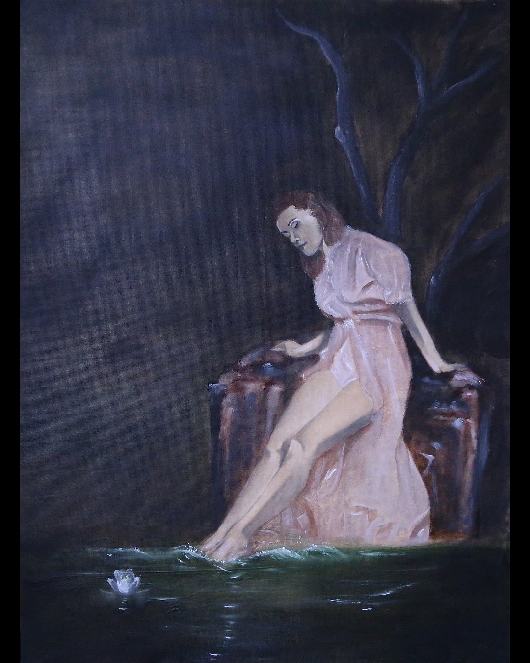 A woman with red hair in a pink dress sitting on a rock by the water, touching her feet in the water, with a dark tree in the background.