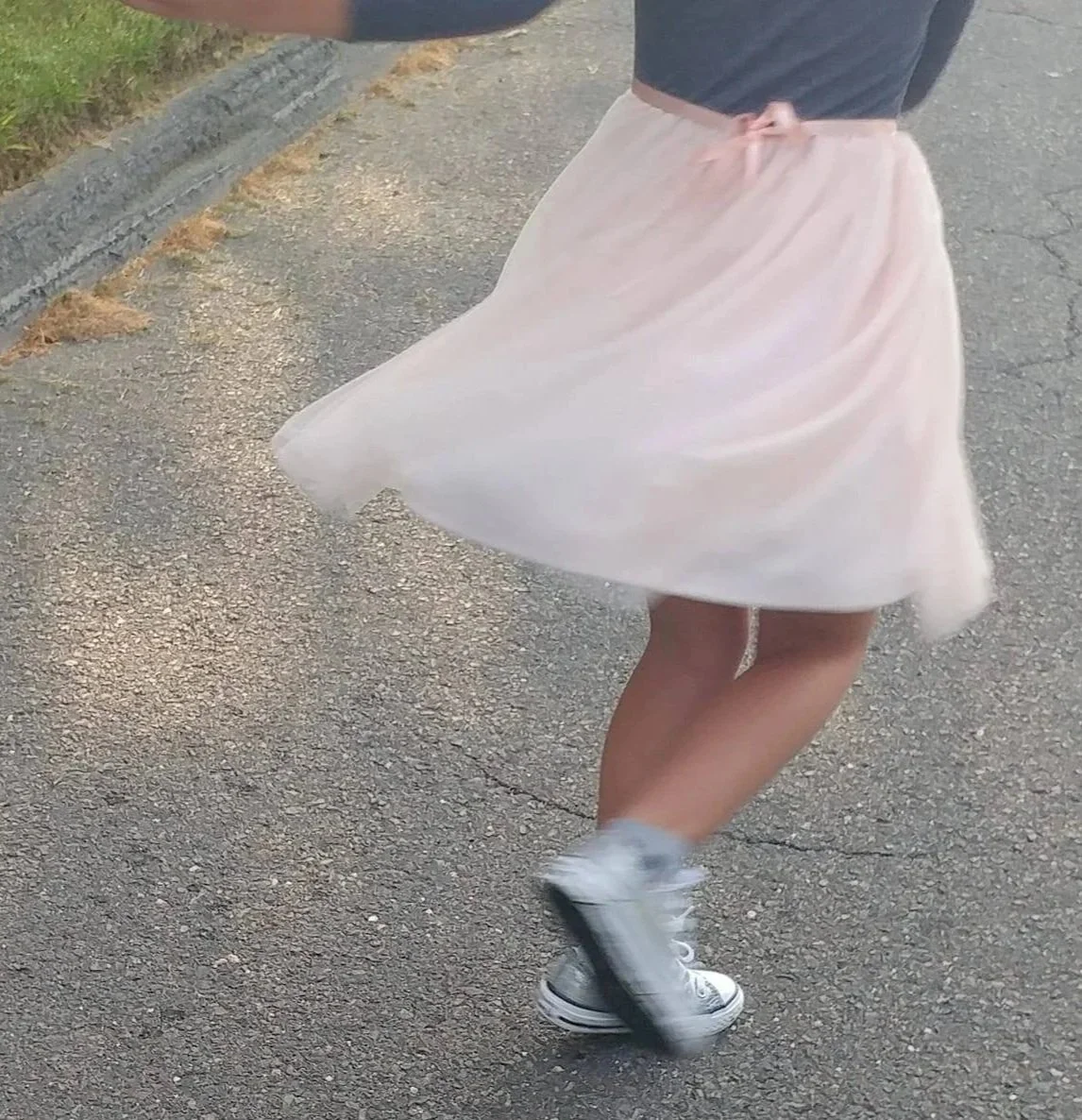 Person wearing a white flowy skirt, black top, and sneakers walking on a paved sidewalk with grass on the side.