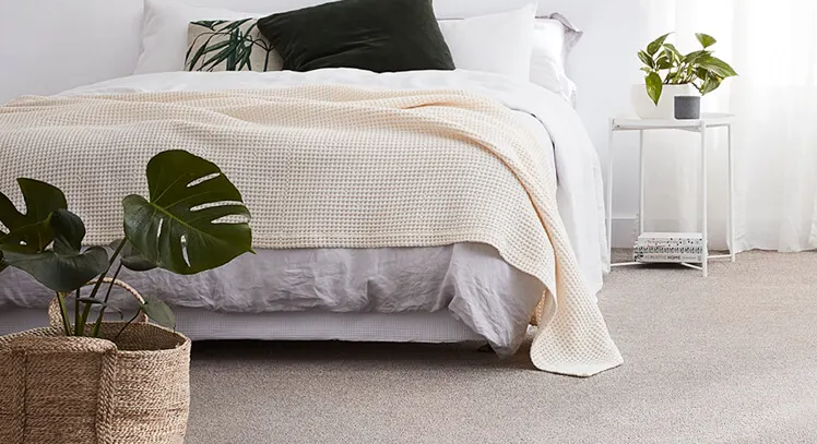 A bedroom with a bed covered in white and cream bedding, a dark pillow, a small white side table with a potted plant, and a large basket with green monstera leaves, all on a light gray carpet.
