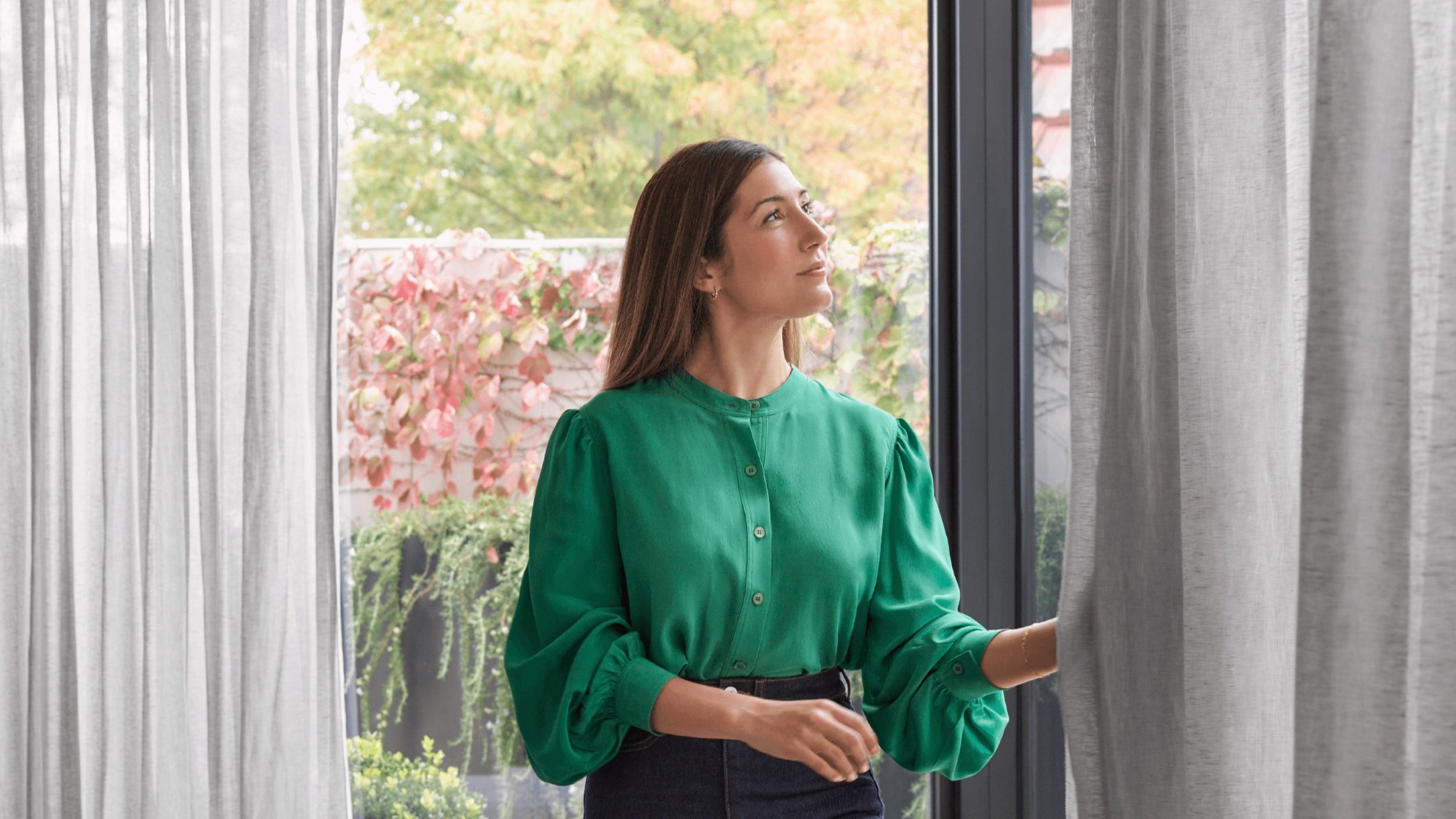 Woman in a green blouse standing by a window and opening gray curtains