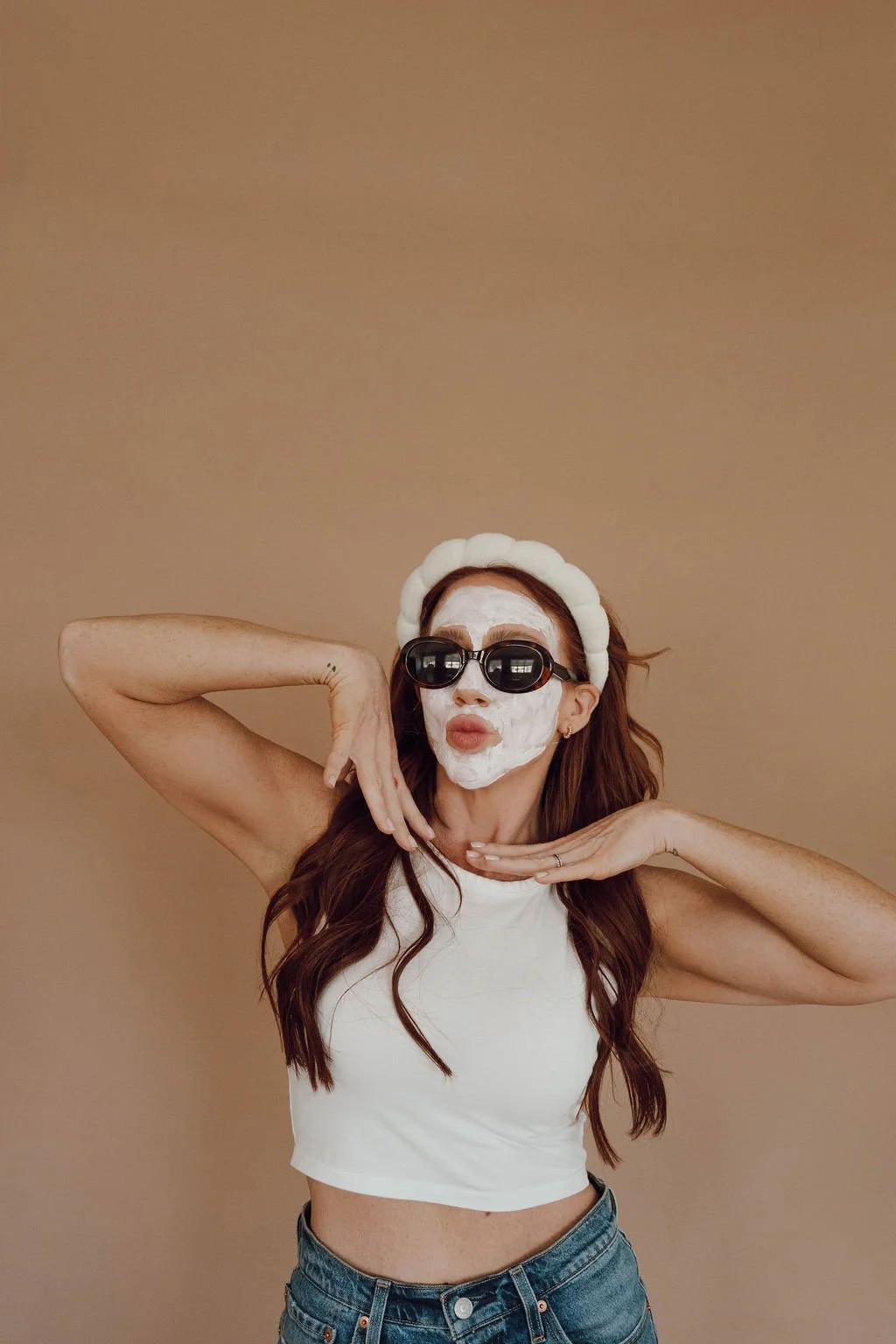A woman with long brown hair wearing a white face mask, black sunglasses, a white headband, a white sleeveless crop top, and blue jeans posing with her hands near her face against a plain beige background.