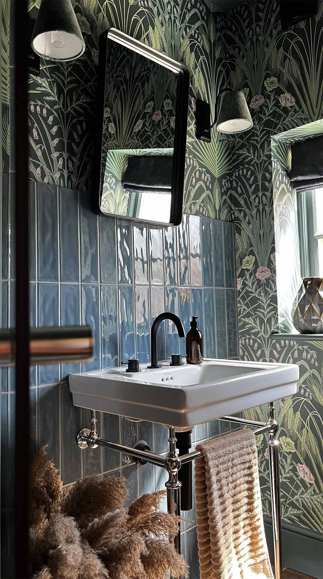 A vintage-style bathroom with green botanical wallpaper, a white sink with black fixtures, and a large mirror. Brown towels hang on a chrome towel rack, with a brown bottle on the sink. Dried pampas grass in the corner adds a natural touch. Two wall-mounted lamps and a window allow natural light.
