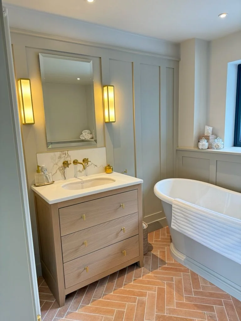 Modern bathroom with a white freestanding bathtub, a wooden vanity with a marble countertop, yellow accent lighting, and a window with decorative jars on the ledge.