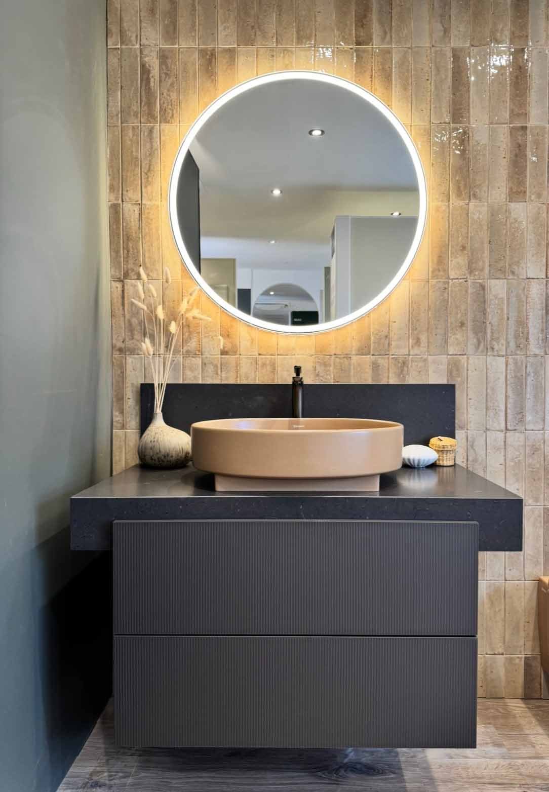 terracotta round basin on a black fluted vanity unit with an illuminated round mirror above on a brown tiled background