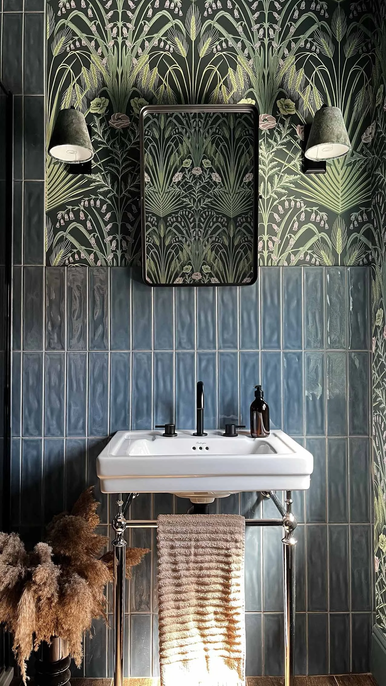 A bathroom with a white sink on a chrome stand, a beige towel hanging, a soap dispenser, dark green patterned wallpaper with floral and leaf designs, blue tiles behind the sink, two wall lamps, a mirror, and dried plants in a black vase.