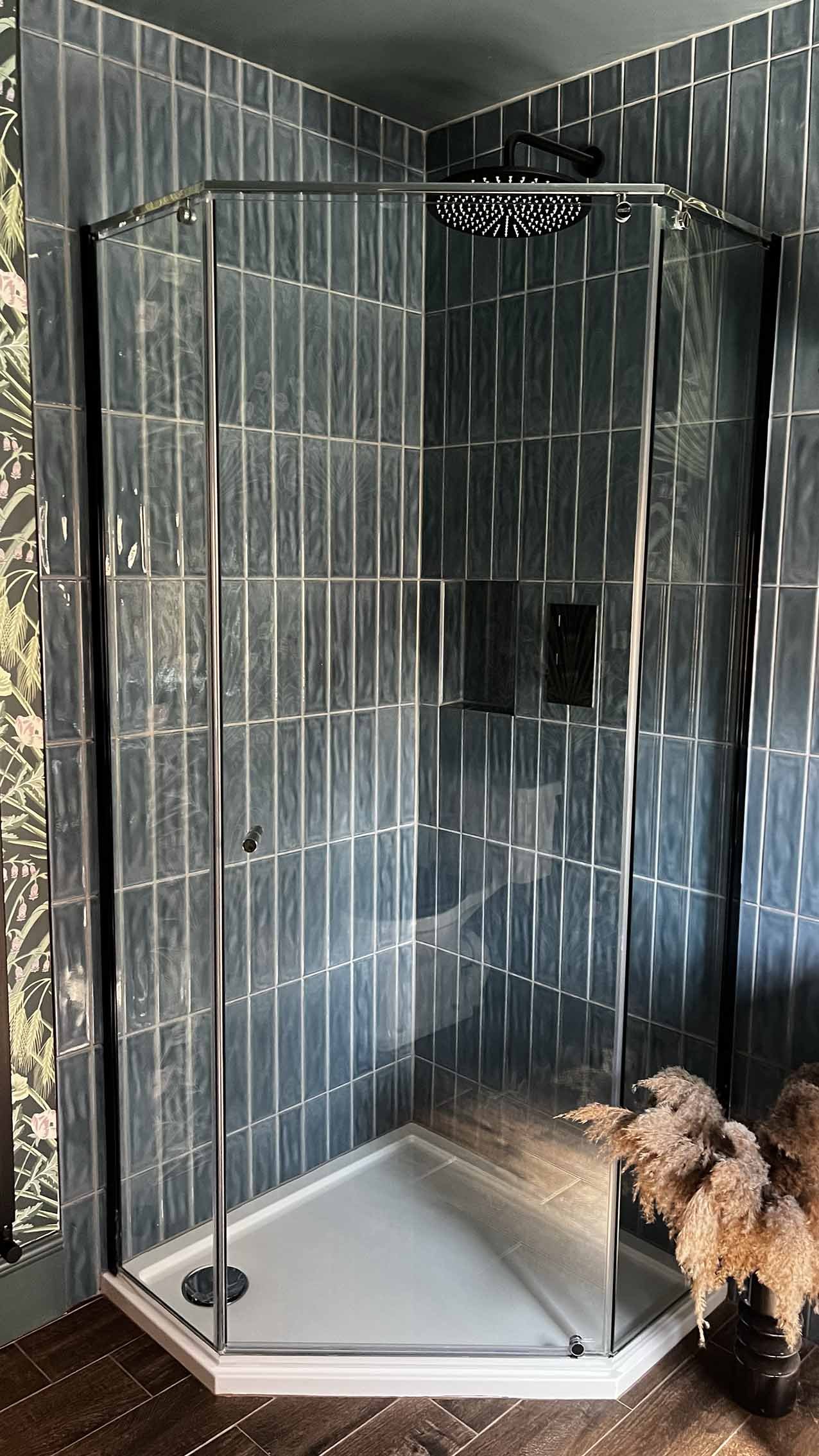 A glass shower enclosure with metal framing in a bathroom with greenish-blue tiled walls and a dark tile floor. There is a decorative floral wallpaper on the left side and a plant with dried flowers on the right in a black pot. The shower has a black overhead rain showerhead.