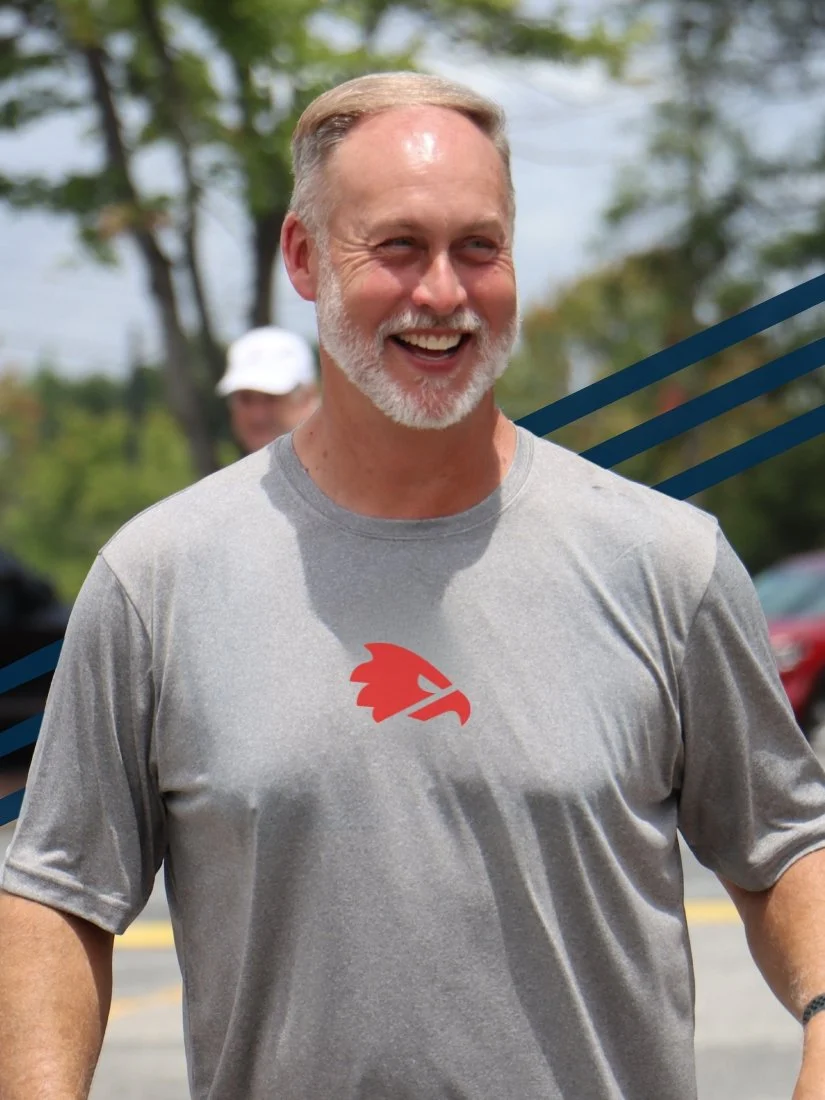 A smiling man with gray hair and a beard wearing a gray t-shirt with a red logo, outdoors on a sunny day.