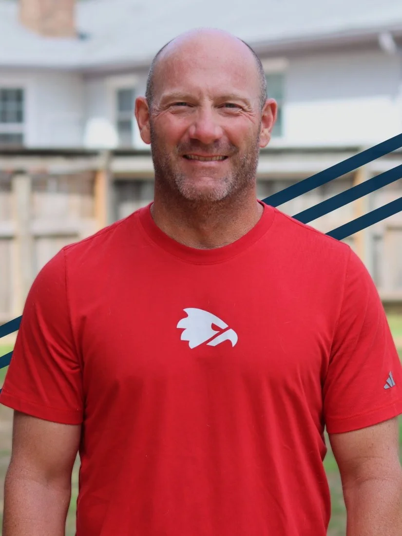 A man with a shaved head and beard, wearing a red sports shirt with a white logo of a stylized eagle head, standing outdoors in a backyard with a wooden fence and houses in the background.