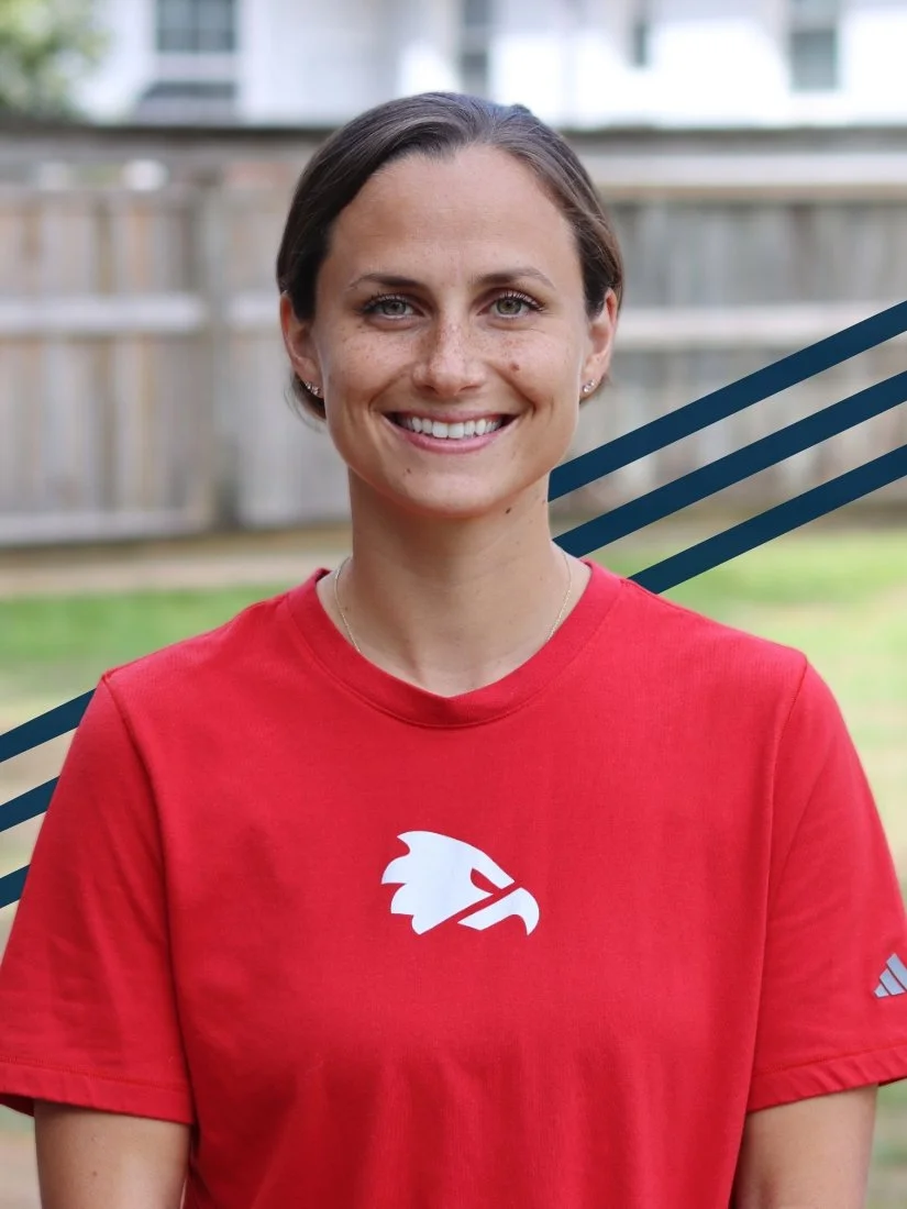 Smiling woman with short brown hair wearing a red Adidas shirt with a white eagle logo, standing outdoors in a backyard with a wooden fence and green grass.