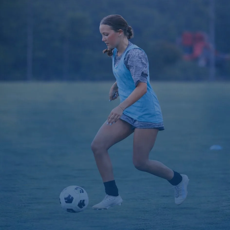 A young woman in athletic clothing is playing soccer on a field in the evening or early morning.
