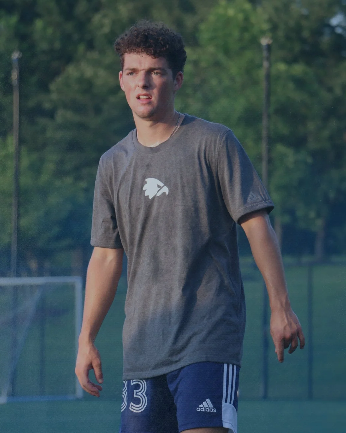 Young man with curly hair in a gray Adidas t-shirt and blue Adidas shorts standing on a soccer field outdoors.