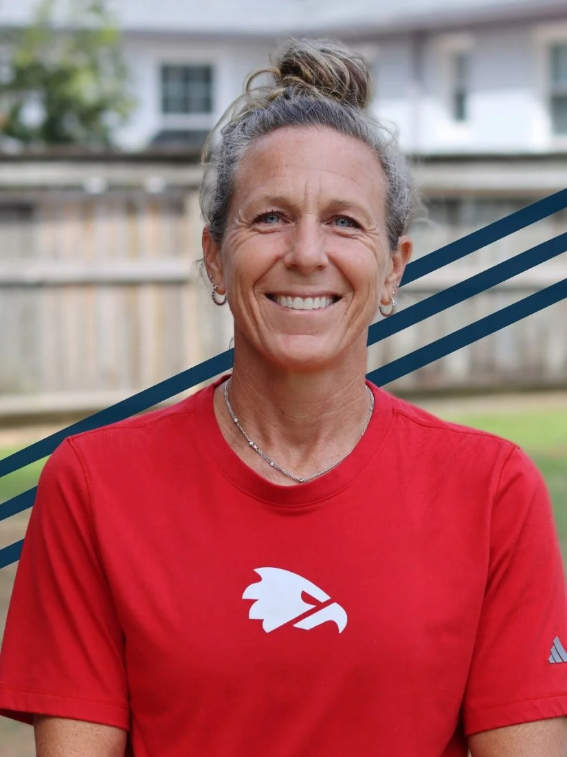 A smiling woman with gray hair tied in a bun, wearing a red sports shirt with a white logo of a stylized head of a Native American, outdoors in a backyard with a wooden fence and houses in the background.