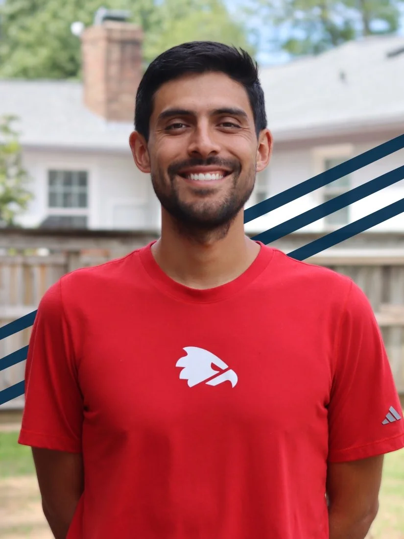 A man standing outdoors in a residential backyard, smiling, wearing a red athletic shirt with a logo of a stylized eagle head. There are houses and trees in the background.