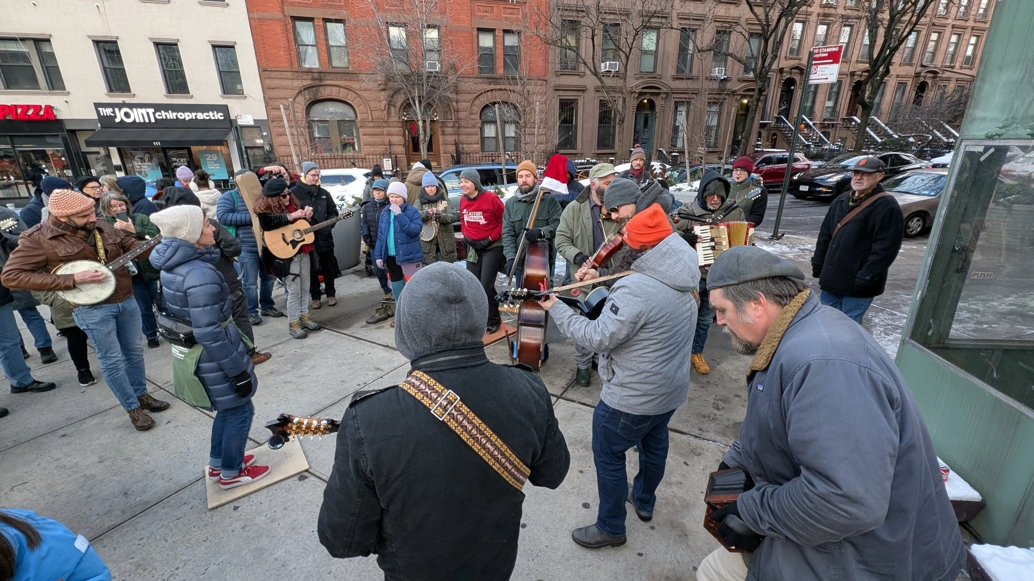 A group of street musicians and spectators gather in a city setting. The musicians play various instruments like a banjo, guitar, violin, and accordion. People are dressed in winter clothing, and the scene is lively with urban buildings and parked ca