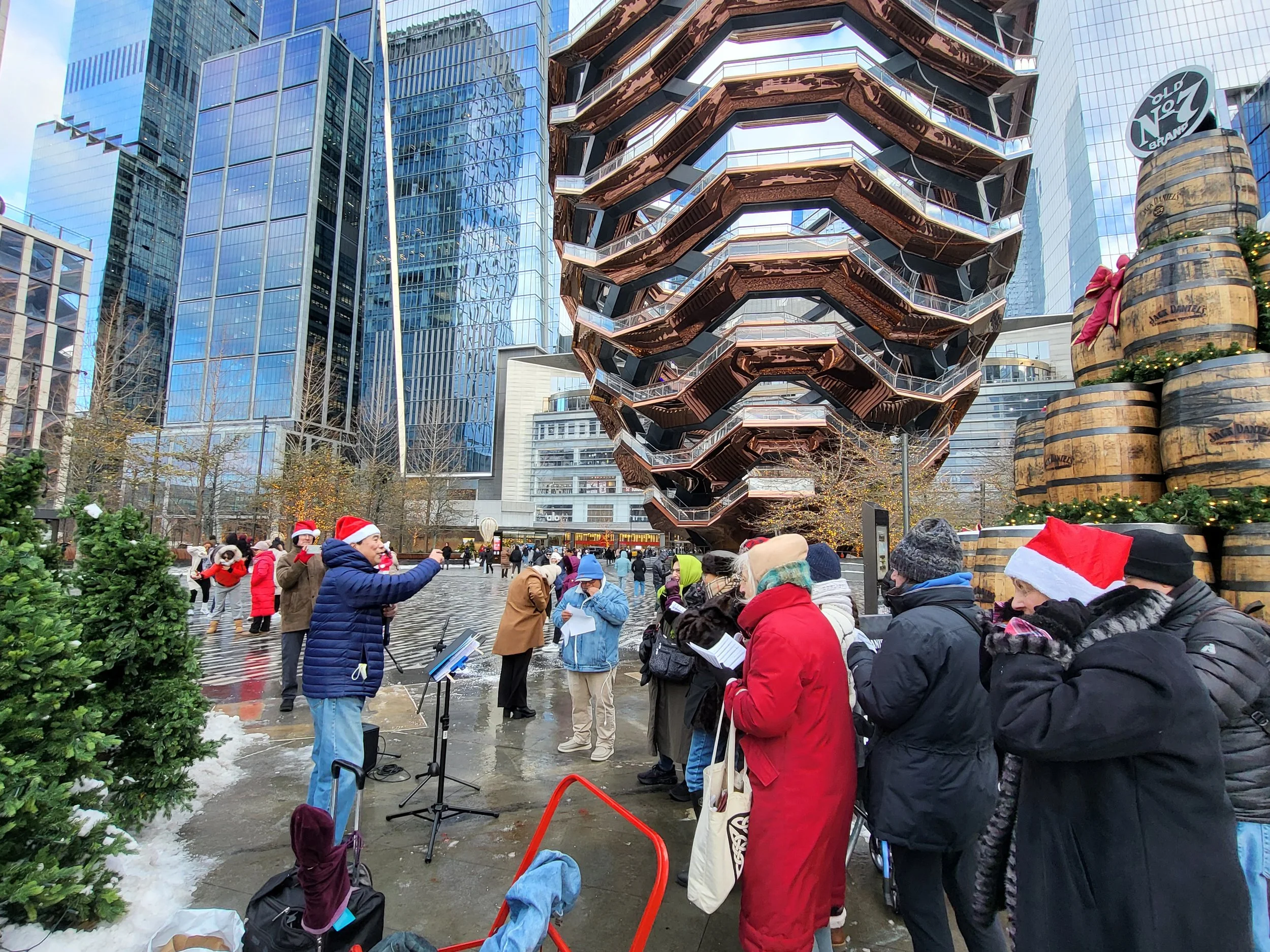 Group of people wearing winter clothing and Santa hats singing outdoors in a city plaza, with the Vessel structure and tall glass buildings in the background. Christmas trees, stacked barrels, and holiday decorations are visible.