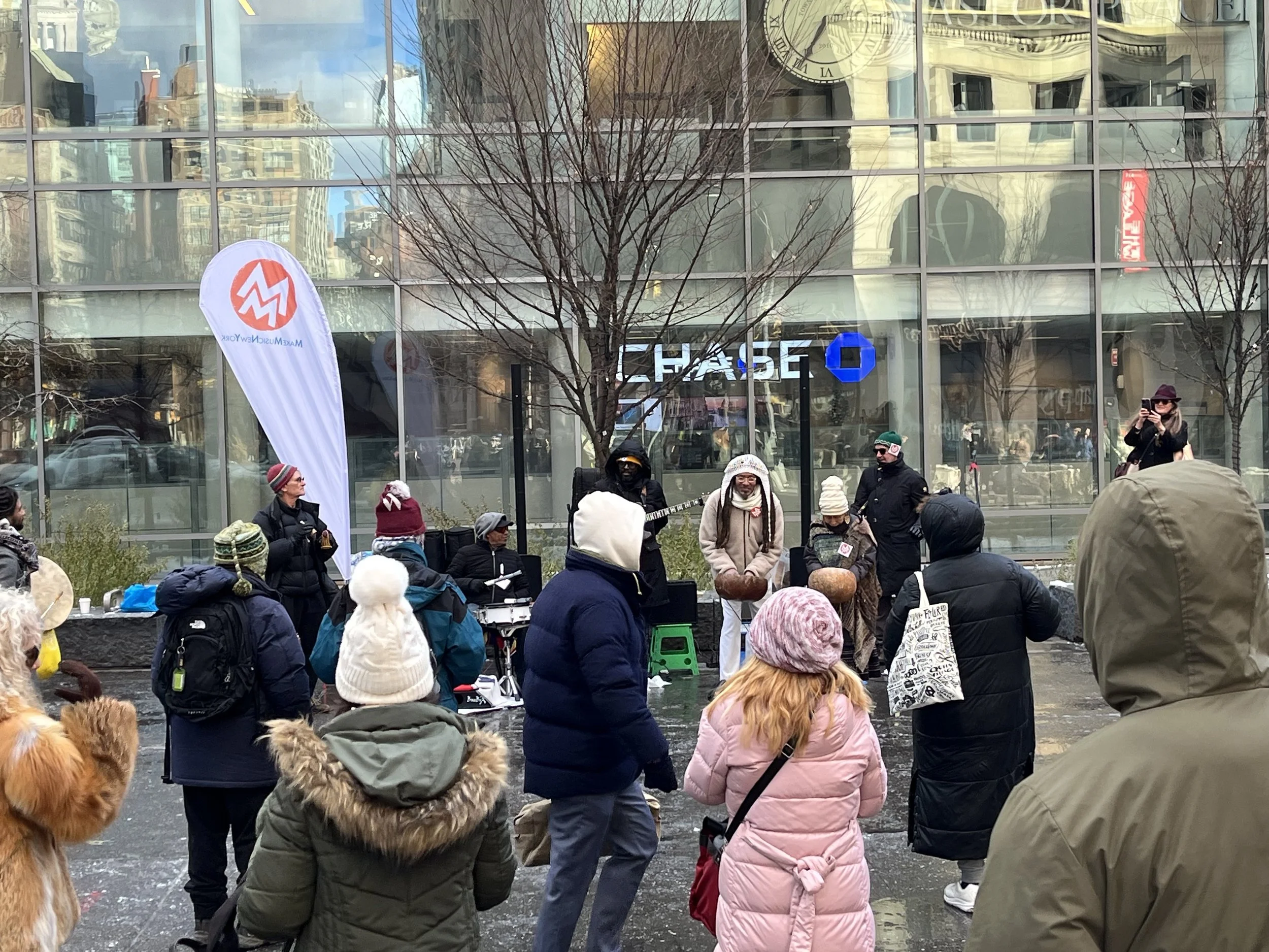 Outdoor street performance with musicians and onlookers in winter clothing outside a Chase bank in an urban area. A large banner with a logo is displayed. Passersby wear coats and hats, indicating cold weather.
