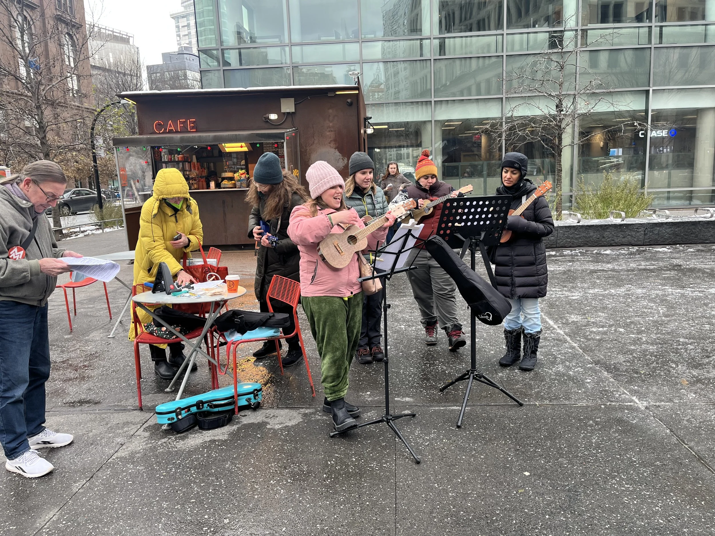 Group of people playing ukuleles and singing outside near a cafe, with music stands and sheet music, in a city setting.