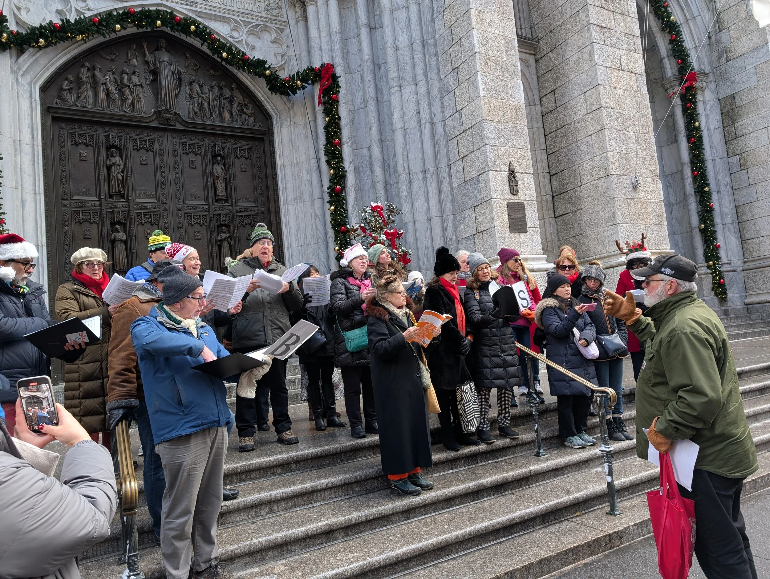 Group of people singing Christmas carols on church steps, wearing winter clothing, in front of a decorated entrance.