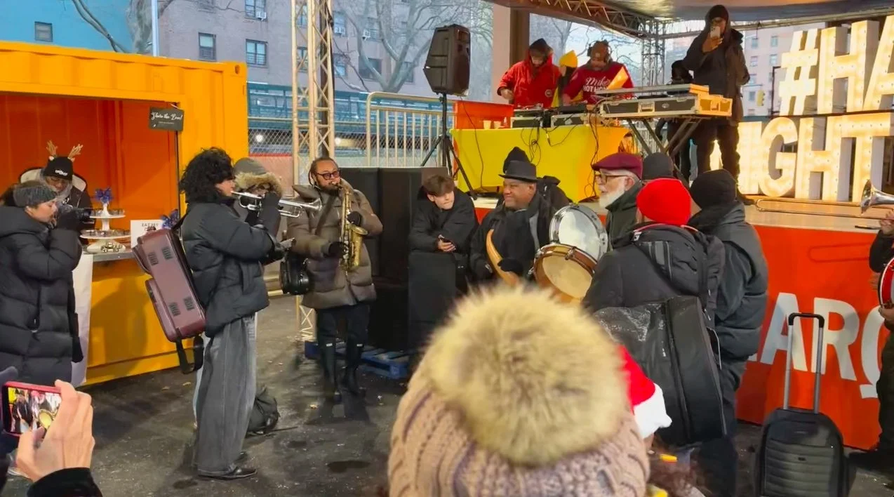 Outdoor event with musicians, DJs, and attendees in winter clothing, next to a bright yellow shipping container and sign with "#Ha...". Band using brass and percussion instruments.