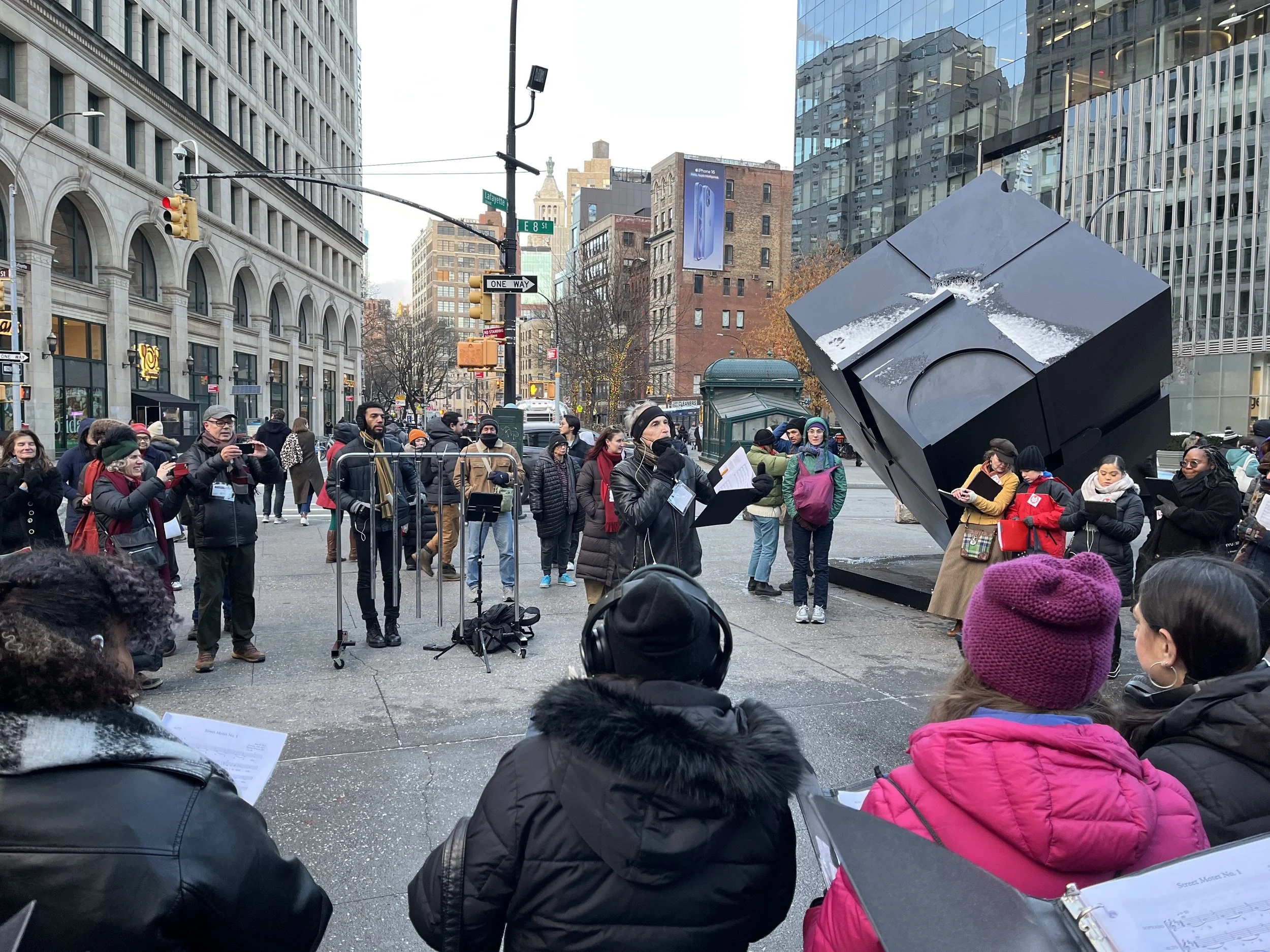 A group of people gathered around the Alamo sculpture in Astor Place, New York City, participating in an outdoor choral performance. The event is taking place in a bustling urban environment, surrounded by buildings and passersby. The participants ar