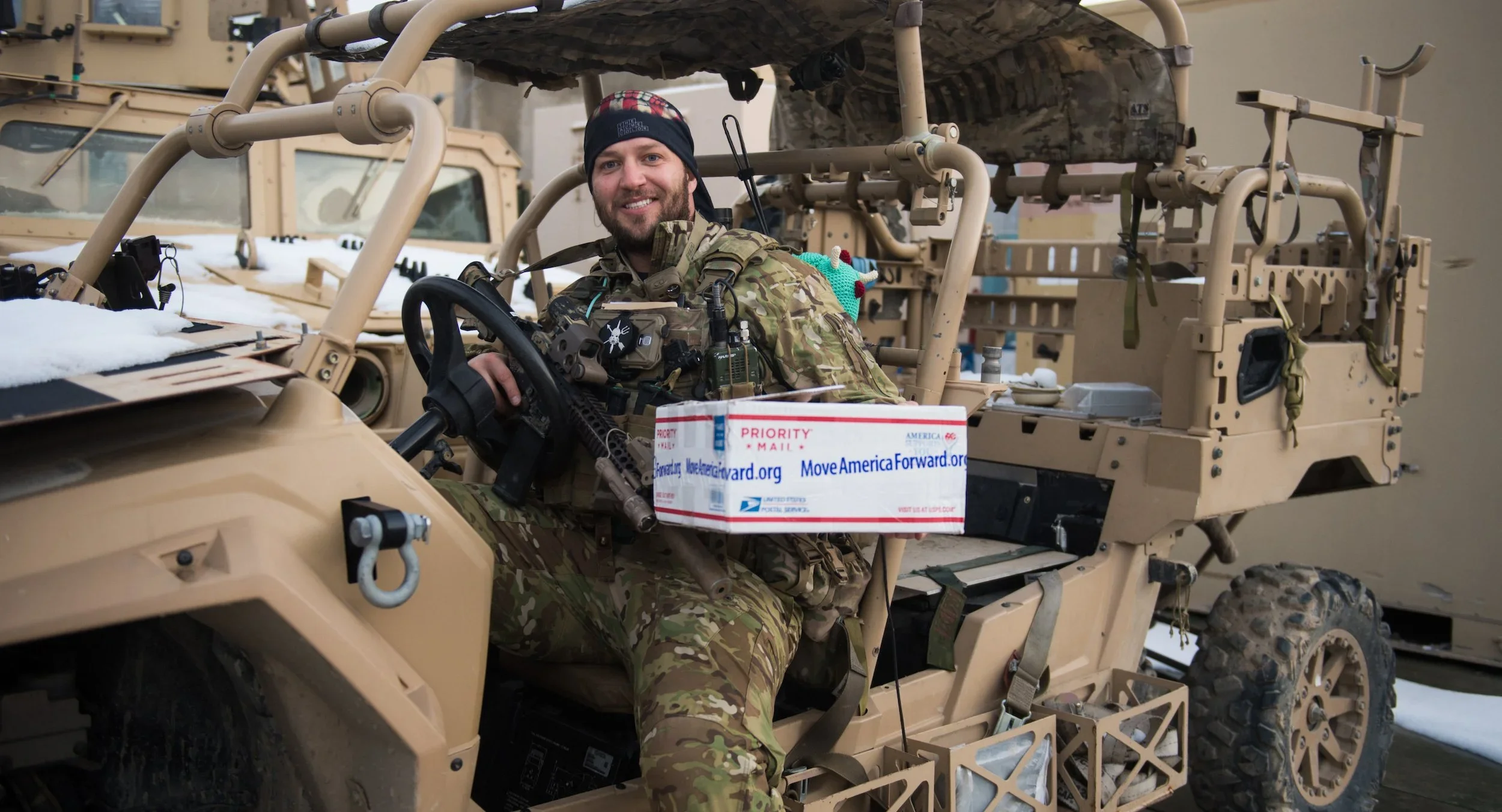 A soldier sitting in a tan military vehicle, smiling, with a box labeled 'Priority Mail' on his lap which is a military care package sent by troopathon that includes candy from candy buyback program, surrounded by military gear and equipment.