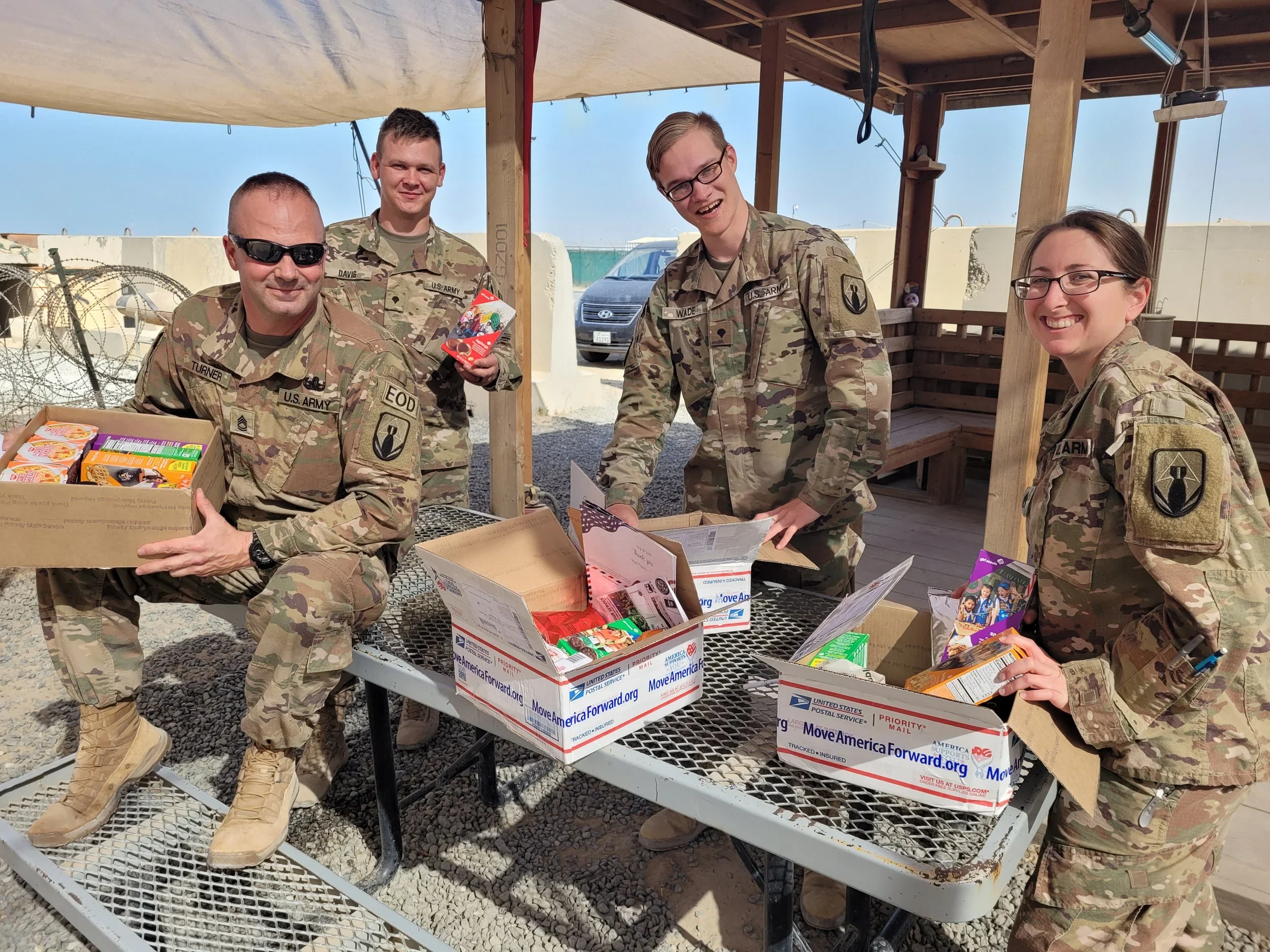 Four U.S. Army soldiers opening military care package boxes of snacks, candy, and supplies from troopathon's candy buyback program outdoors under a shaded wooden structure with a car and a blue sky in the background.