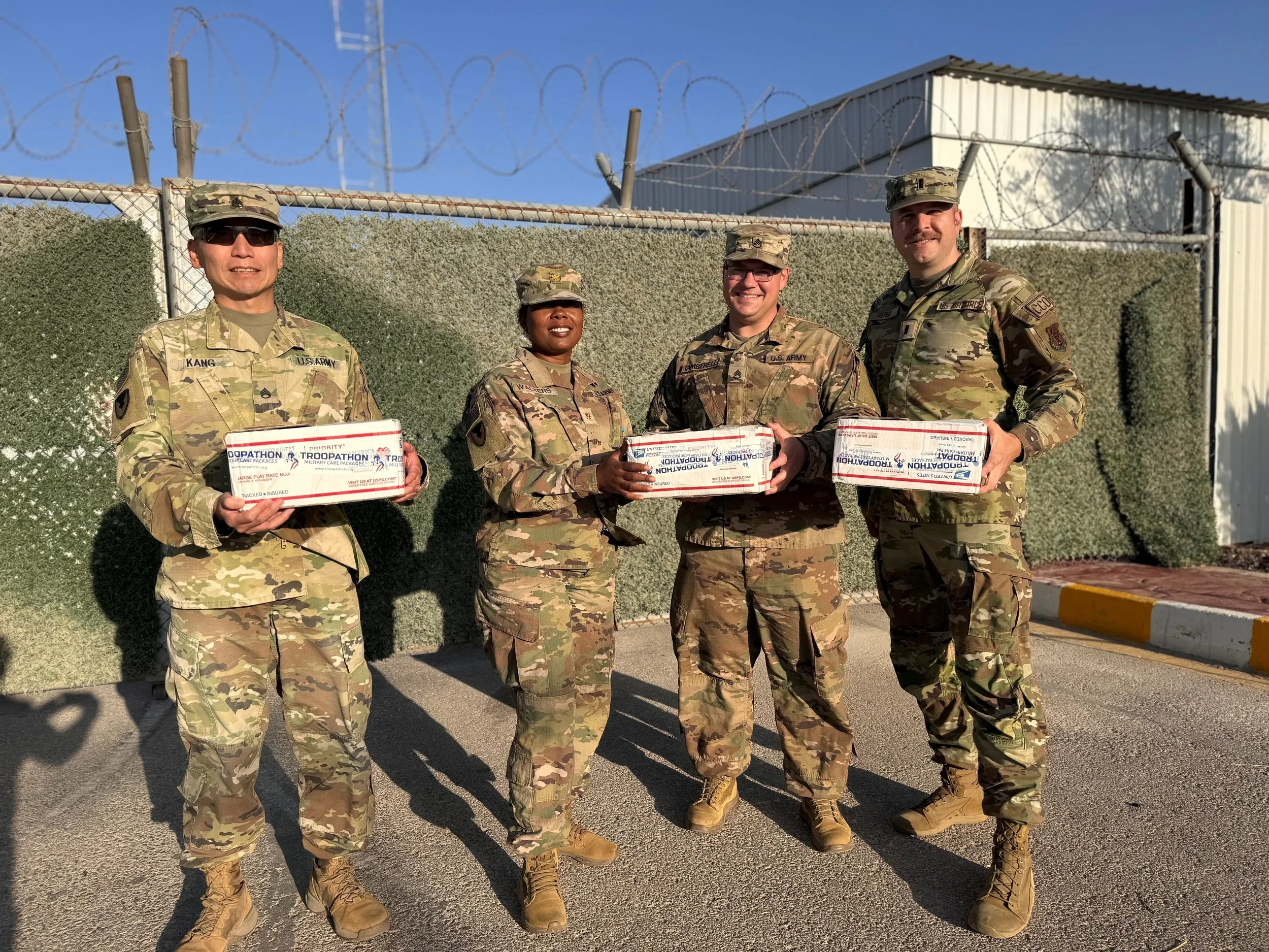 Four soldiers in camouflage uniform standing outdoors holding military care packages with candy from troopathon, smiling, with barbed wire and a building in the background.