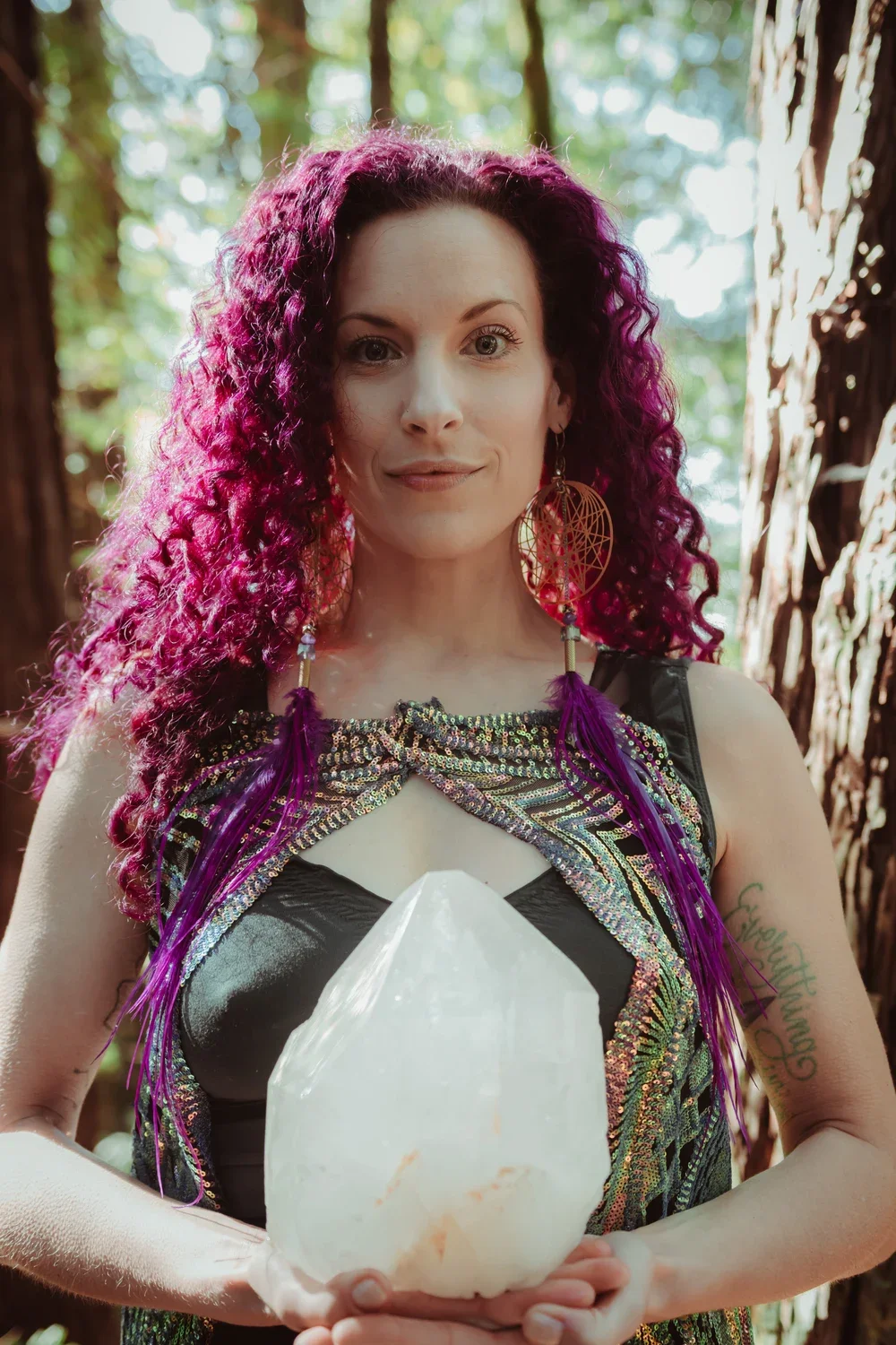 Astra Fox with purple curls and gold earrings, holding a crystal among trees